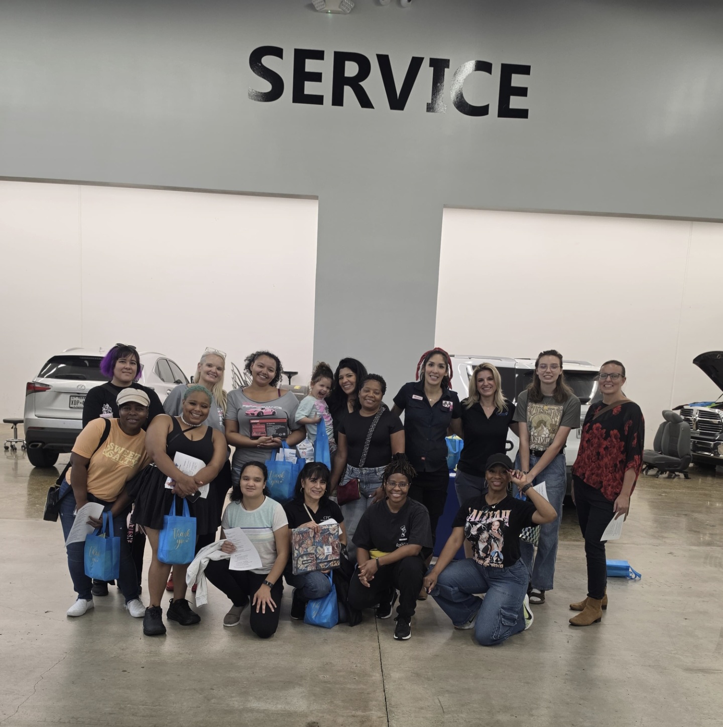 Group of people posing inside a car dealership with a large 'SERVICE' sign above them, some holding blue bags.