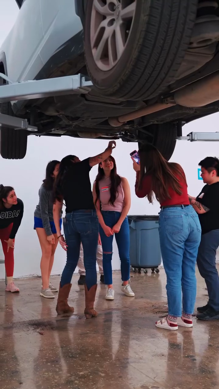 Group of people under a car lifted on a hydraulic lift, discussing and pointing at the vehicle, on a wet surface.