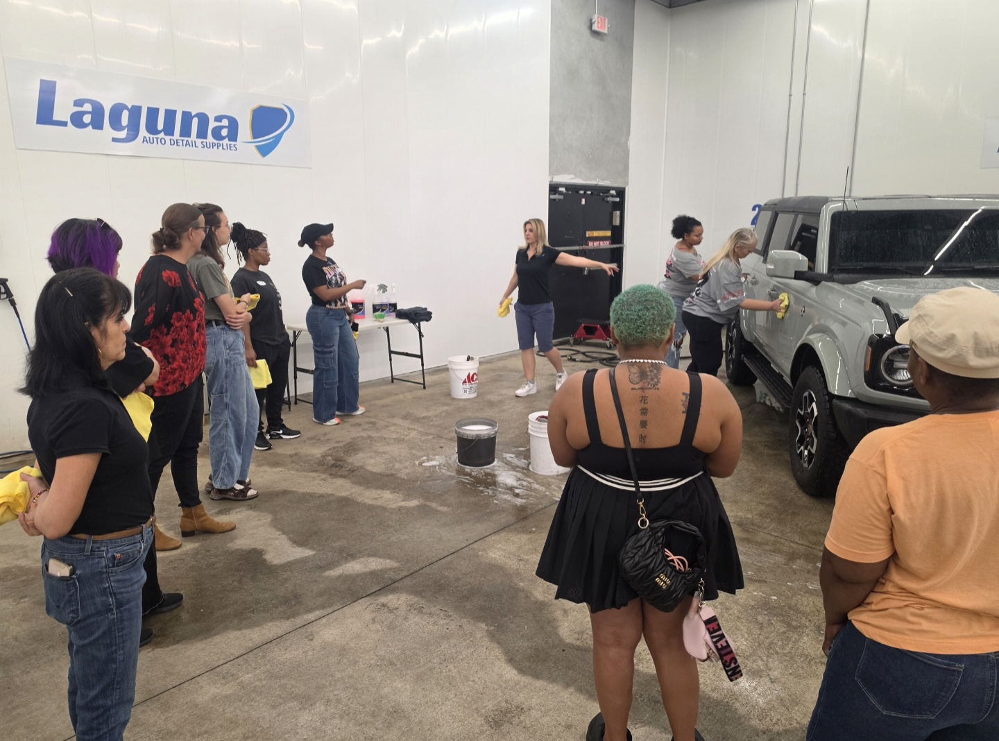 Group of people gathered indoors near vehicles, listening to a woman speaking, with a Laguna sign on the wall.