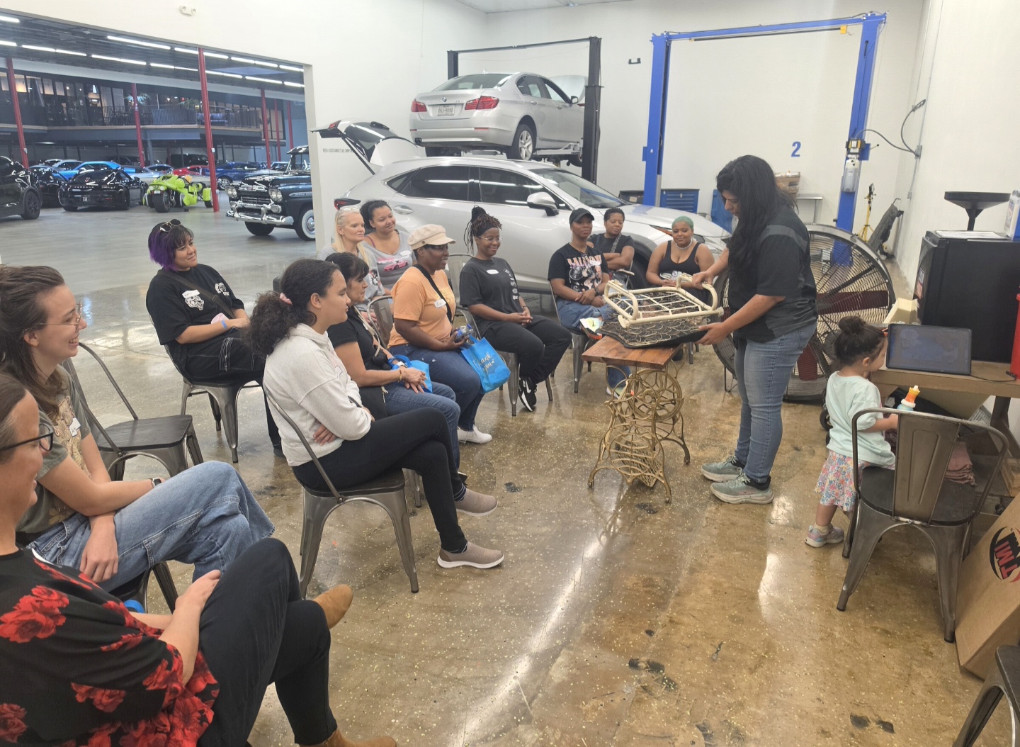 Group of people seated indoors, listening to a woman standing near a table with a cake, in a workshop or garage setting.