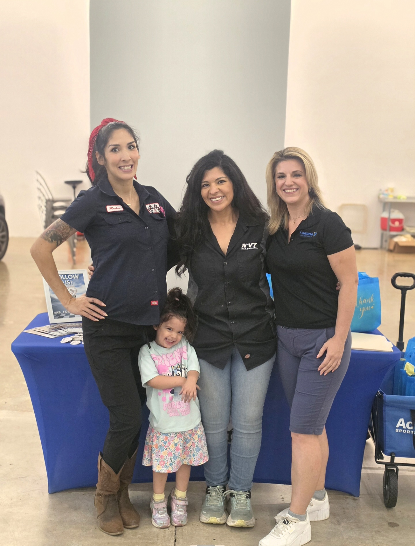 Three women and a young girl standing together indoors, smiling, with a blue table behind them.