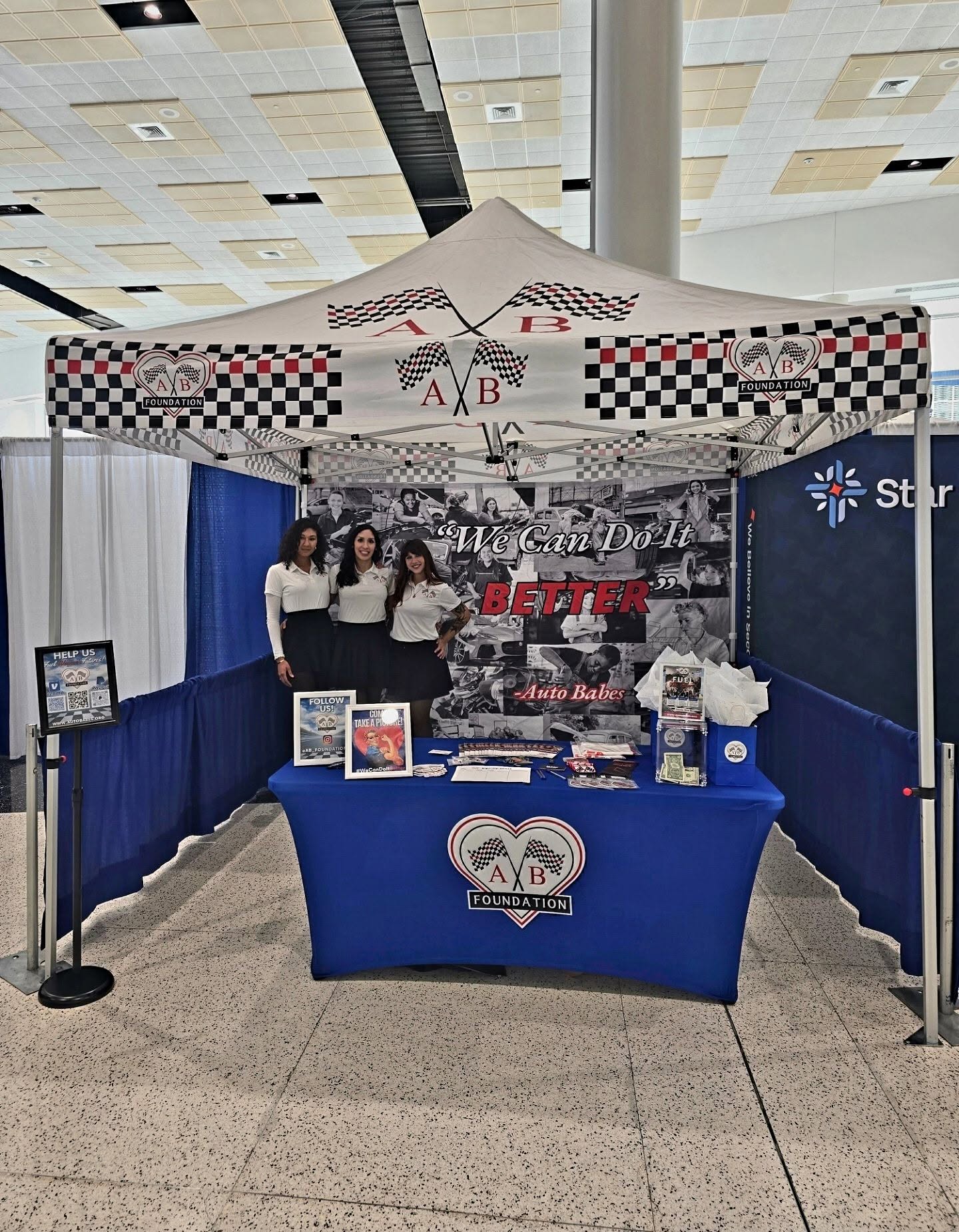 Booth with four people standing behind a table, banners, and photos, inside an indoor space with a high ceiling.