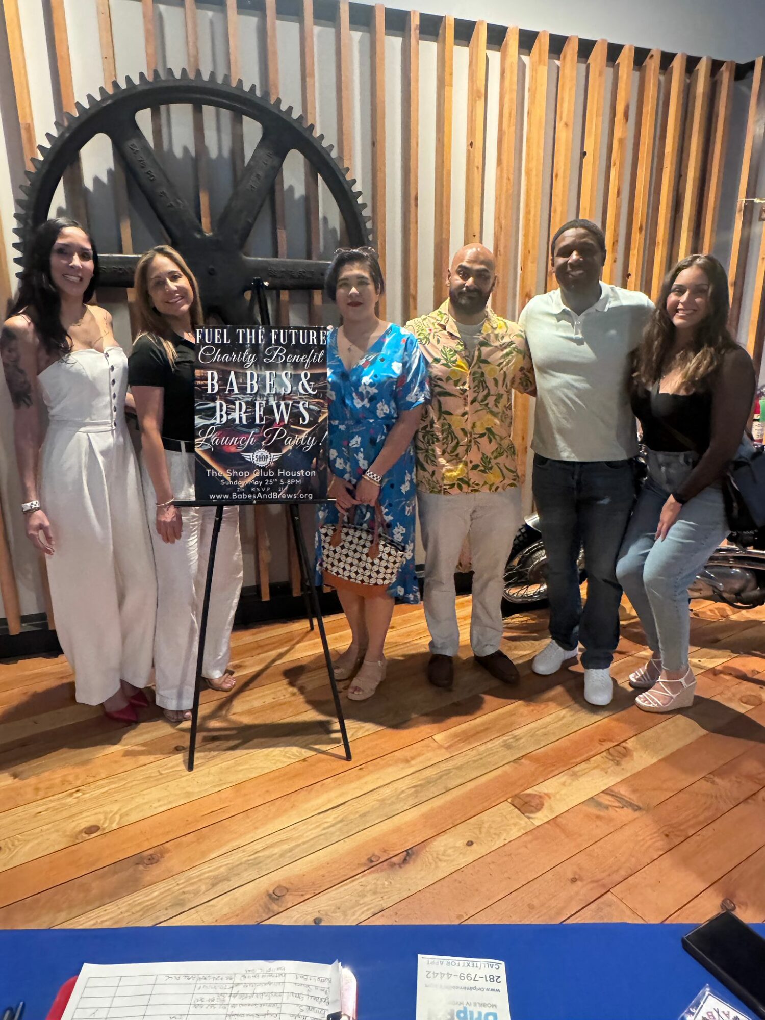 Six people stand in front of a large gear and wooden wall, posing for a photo indoors. A poster on an easel is in front of them.