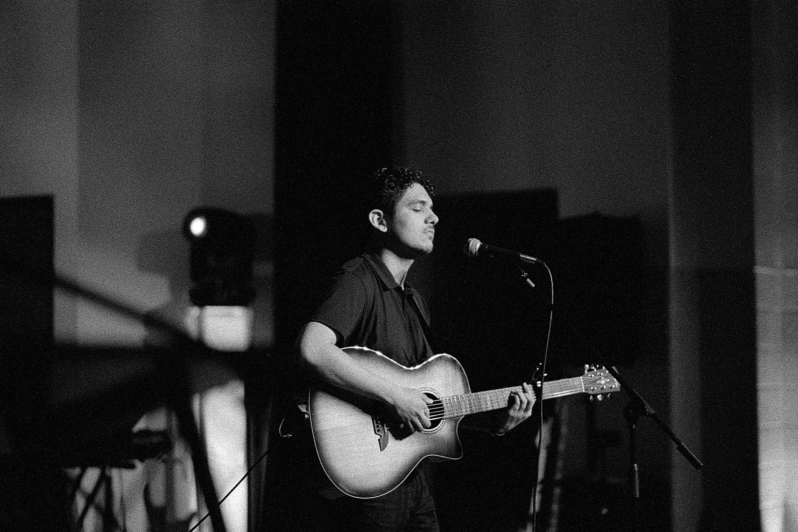 Person playing guitar and singing into a microphone on stage, with a dark background.
