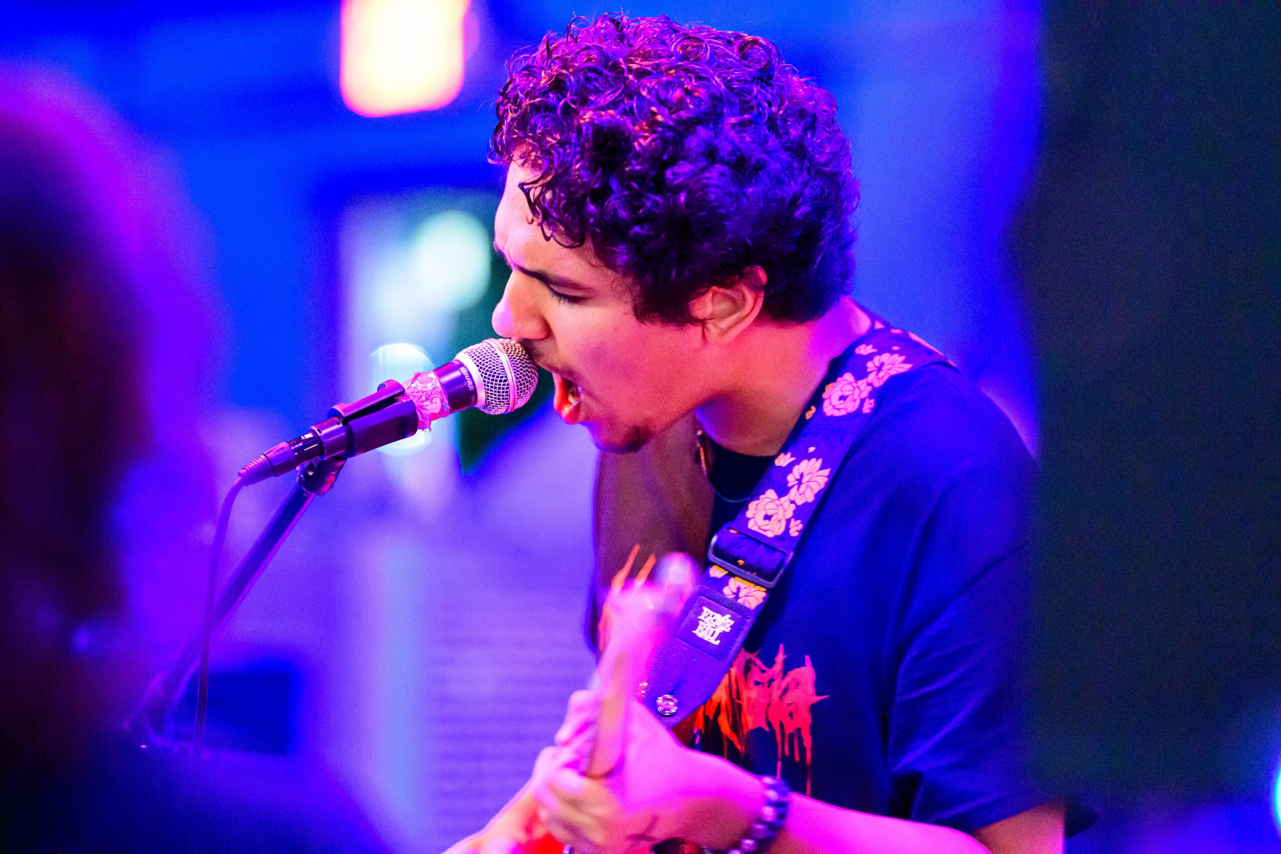 Young person with curly hair singing into a microphone while playing a guitar on stage, with colorful blue and purple lighting.