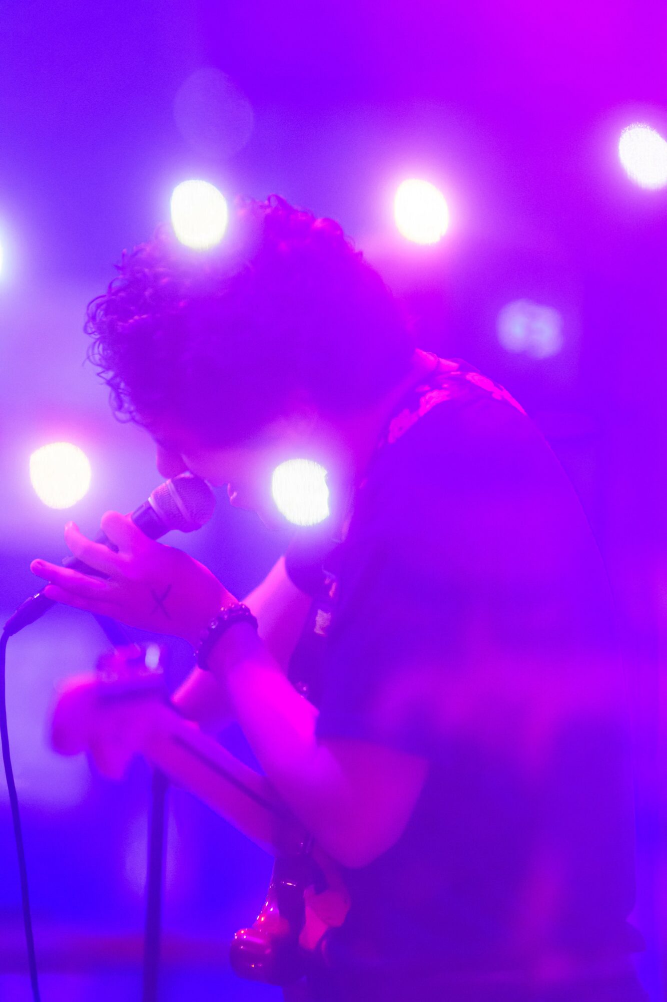 Person holding a microphone on stage with bright purple and pink lights behind them.