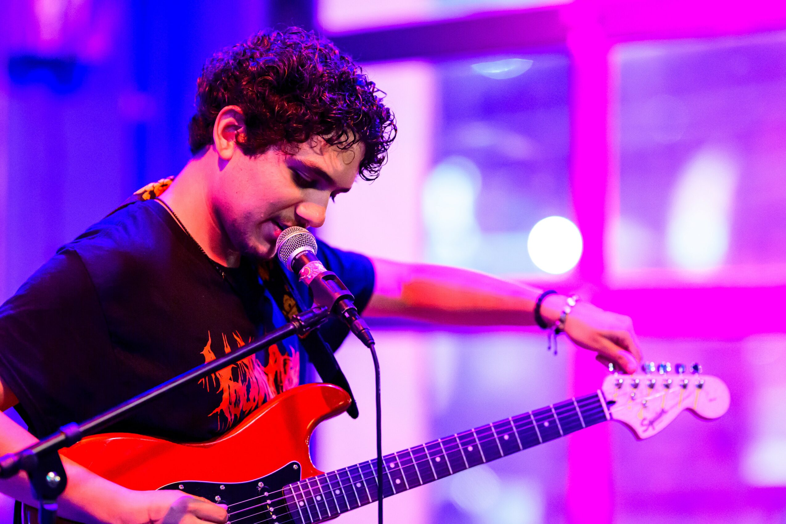 Person with curly hair playing an electric guitar and singing into a microphone, with colorful stage lighting.