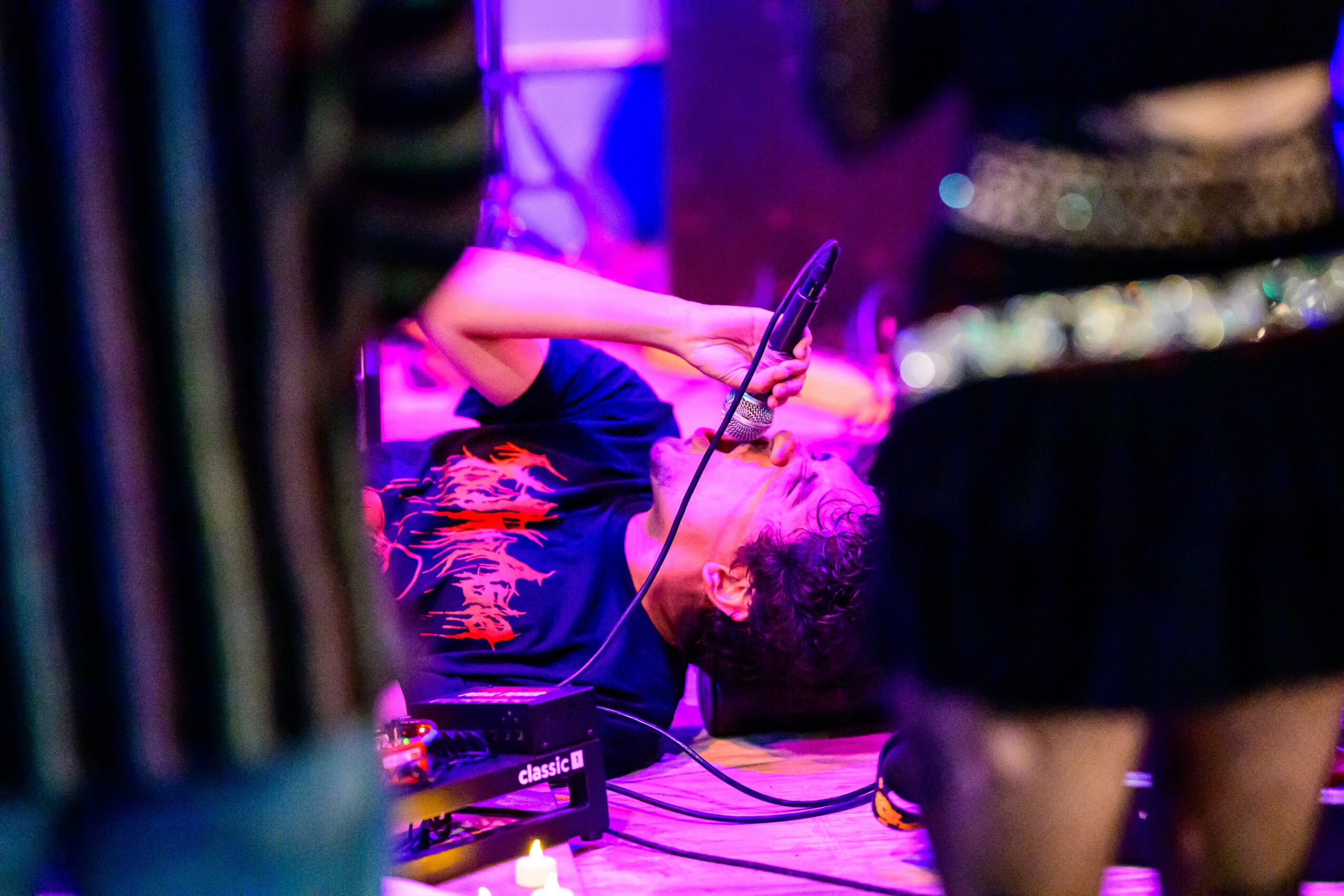 Person singing into a microphone on stage with colorful lighting, wearing a dark shirt with a design, and holding the microphone with one hand.