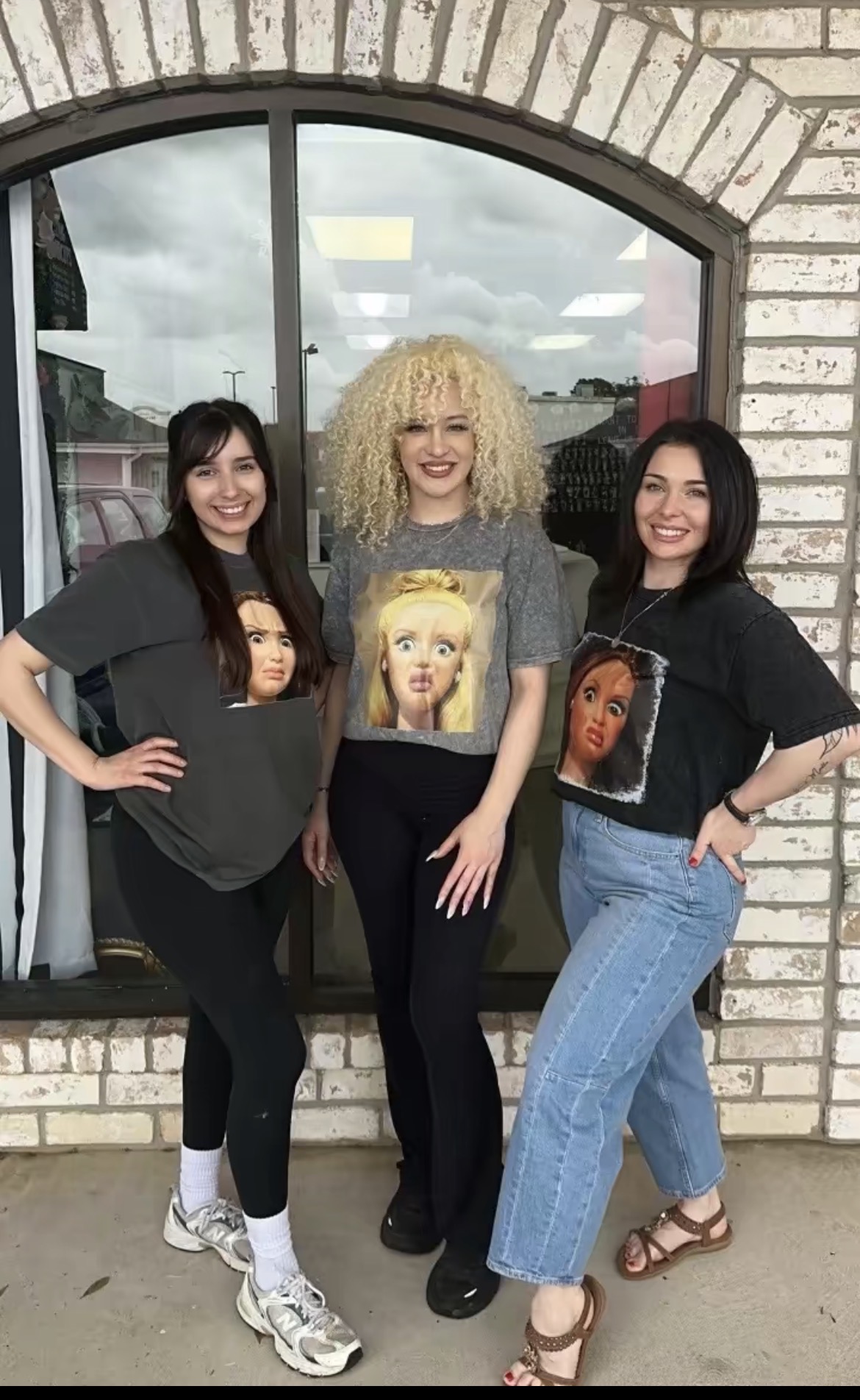 Three women standing in front of a window with a brick wall, smiling, with cloudy sky reflected in the window.