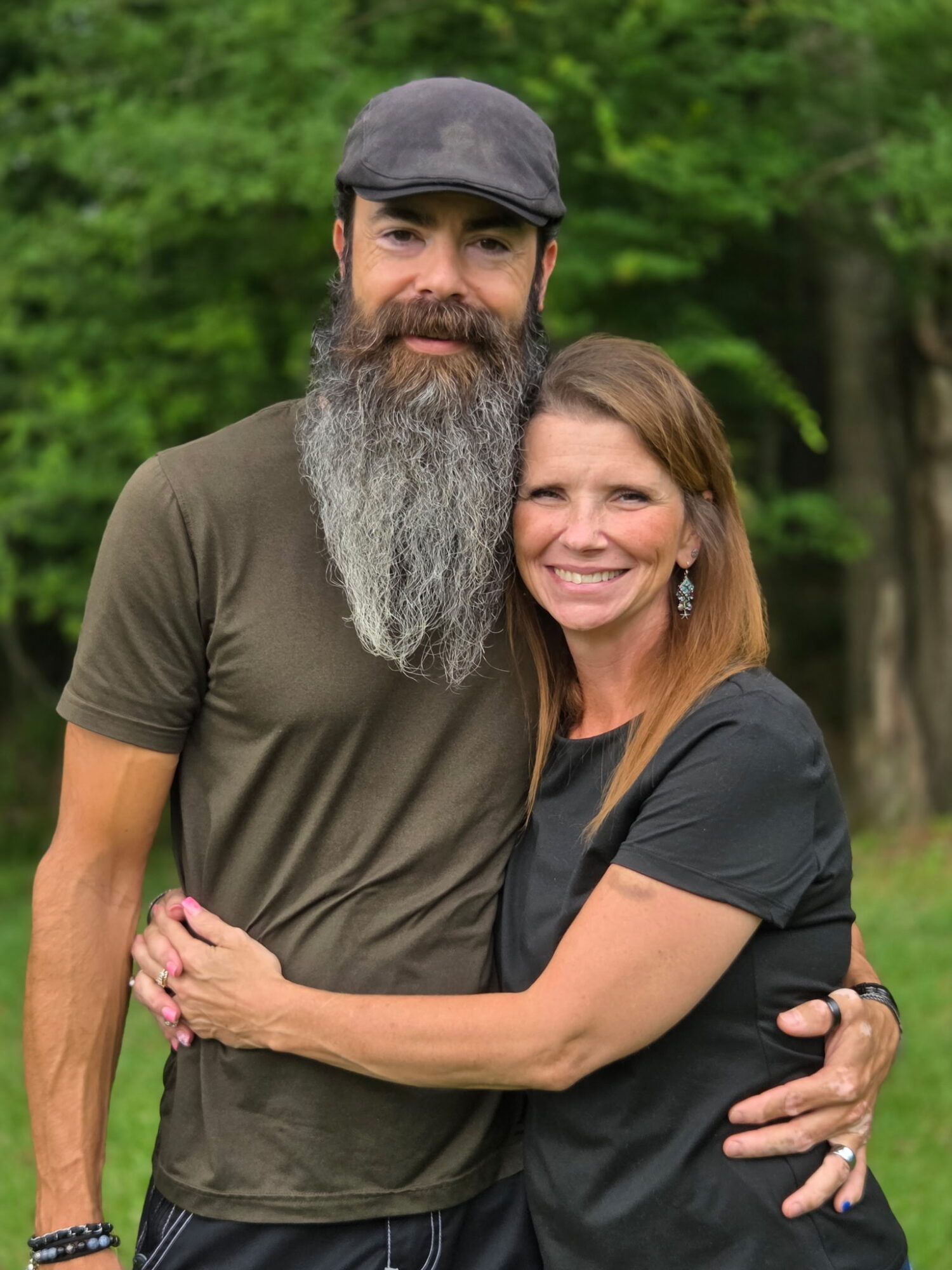 A man with a beard and a woman with shoulder-length hair smiling outdoors, hugging each other.