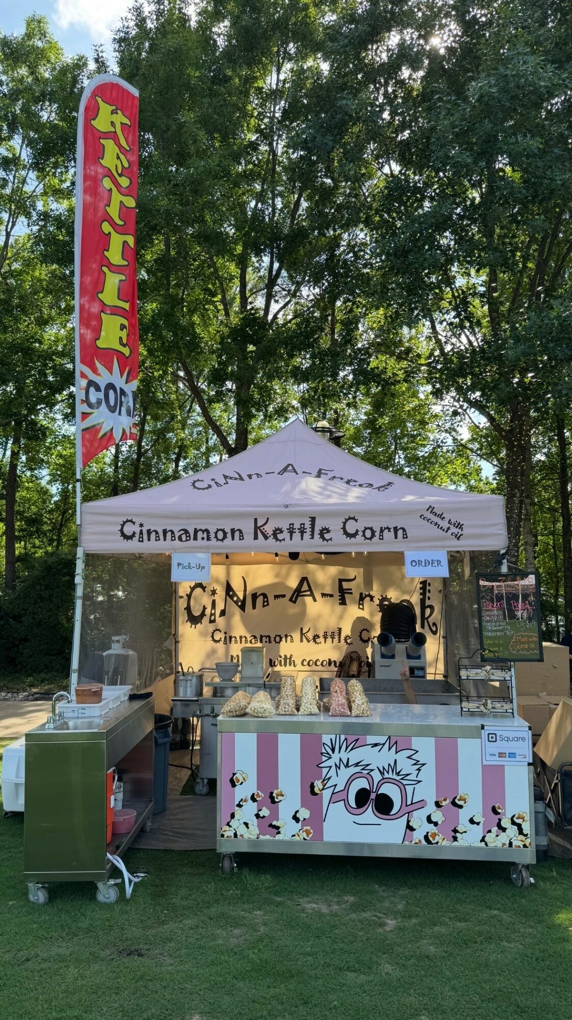 Cinnamon Kettle Corn stand with a white canopy, colorful sign, and a decorated cart outdoors with trees in background.