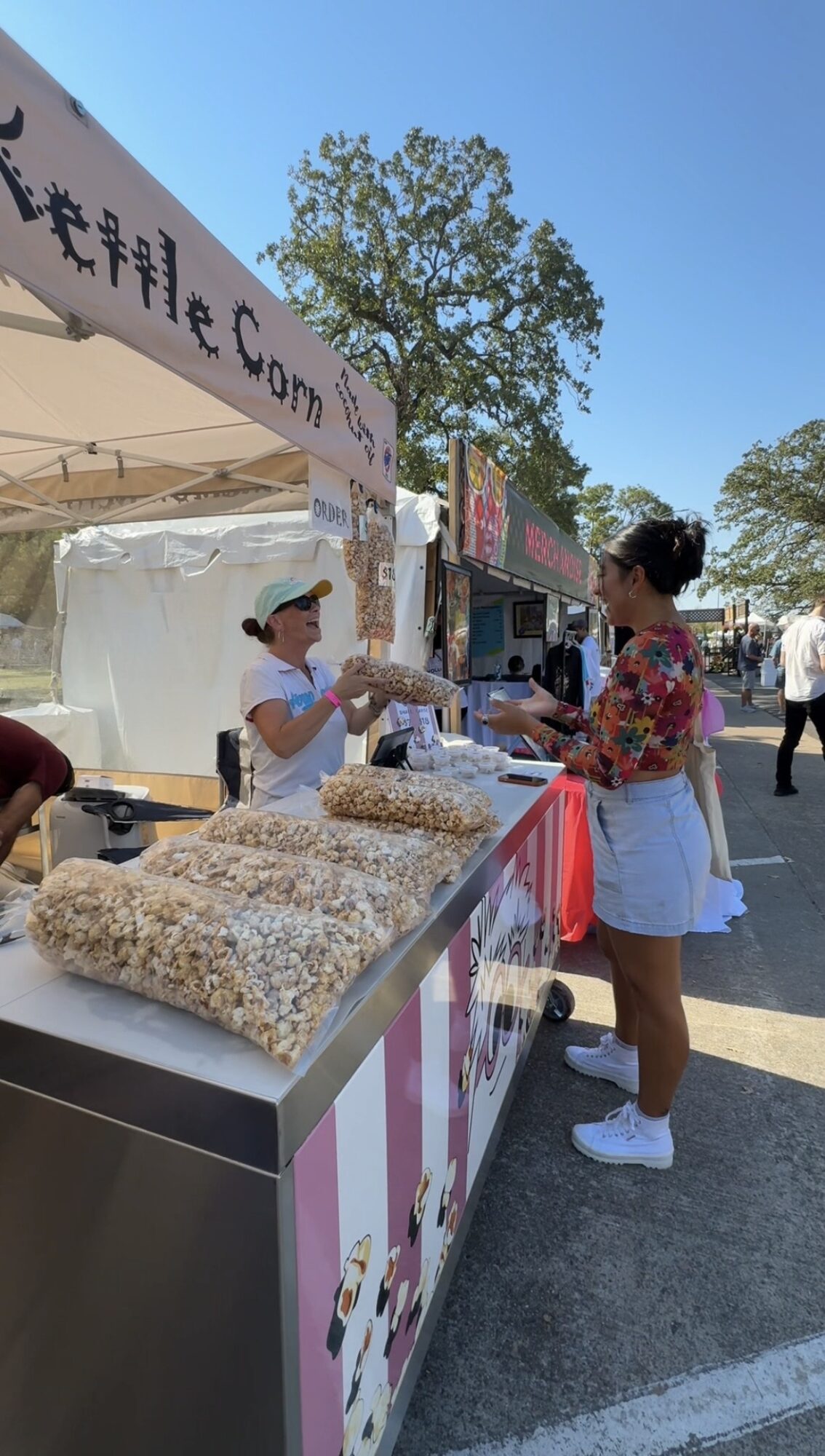 Two women exchange items at a popcorn stand outdoors, with trees and other booths in the background.