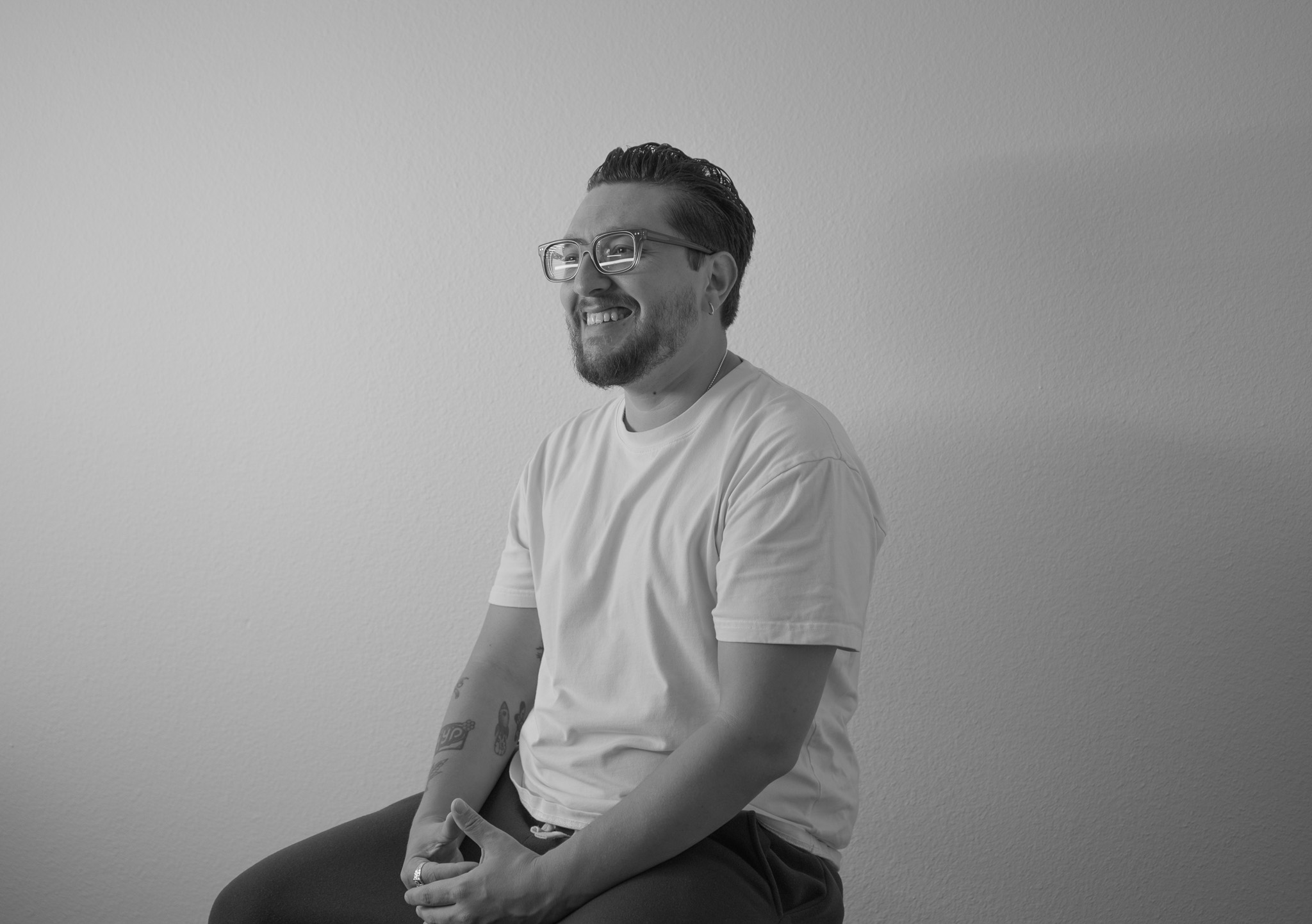 Smiling man with glasses and a beard, sitting against a plain wall, wearing a white t-shirt.