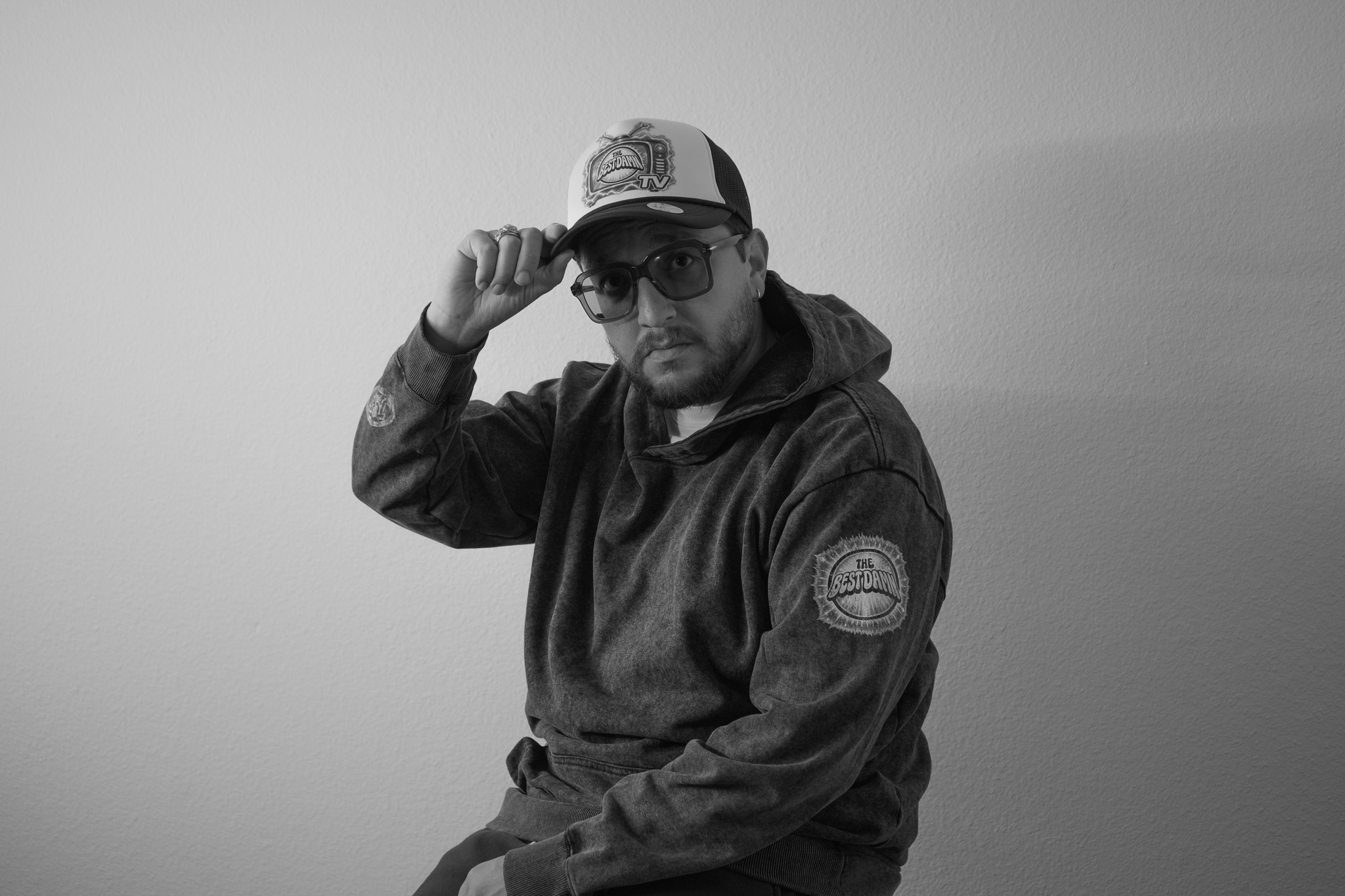 Man wearing sunglasses, baseball cap, and hoodie, adjusting cap with one hand, against plain background.