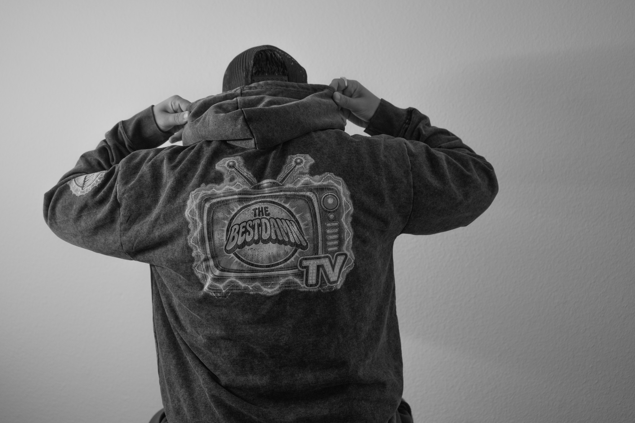 Person adjusting hoodie, back view, wearing a jacket with a logo on the back, plain background.