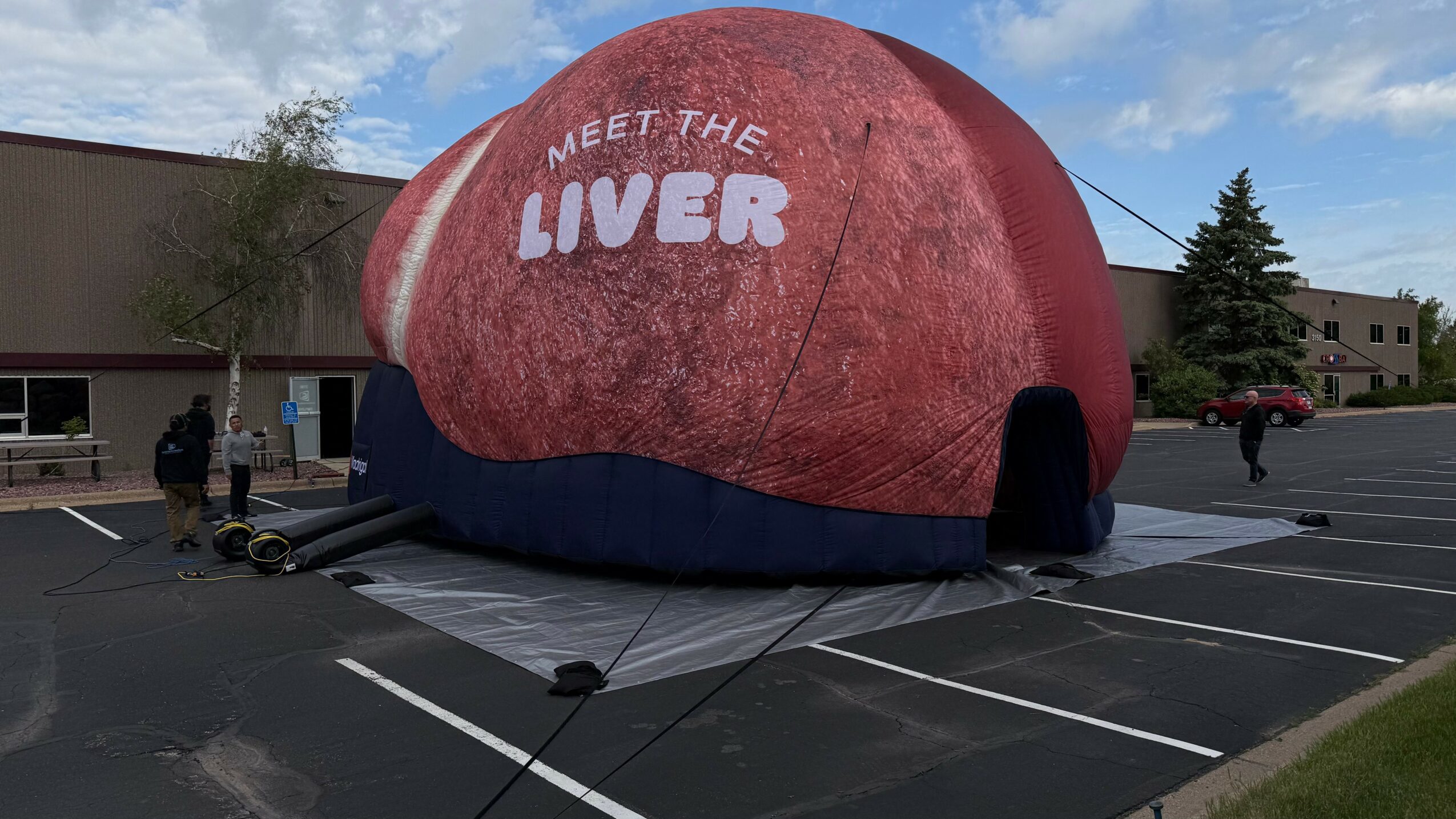 Large inflatable structure resembling a rock with the text 'Meet the Liver' on it, set up in a parking lot.