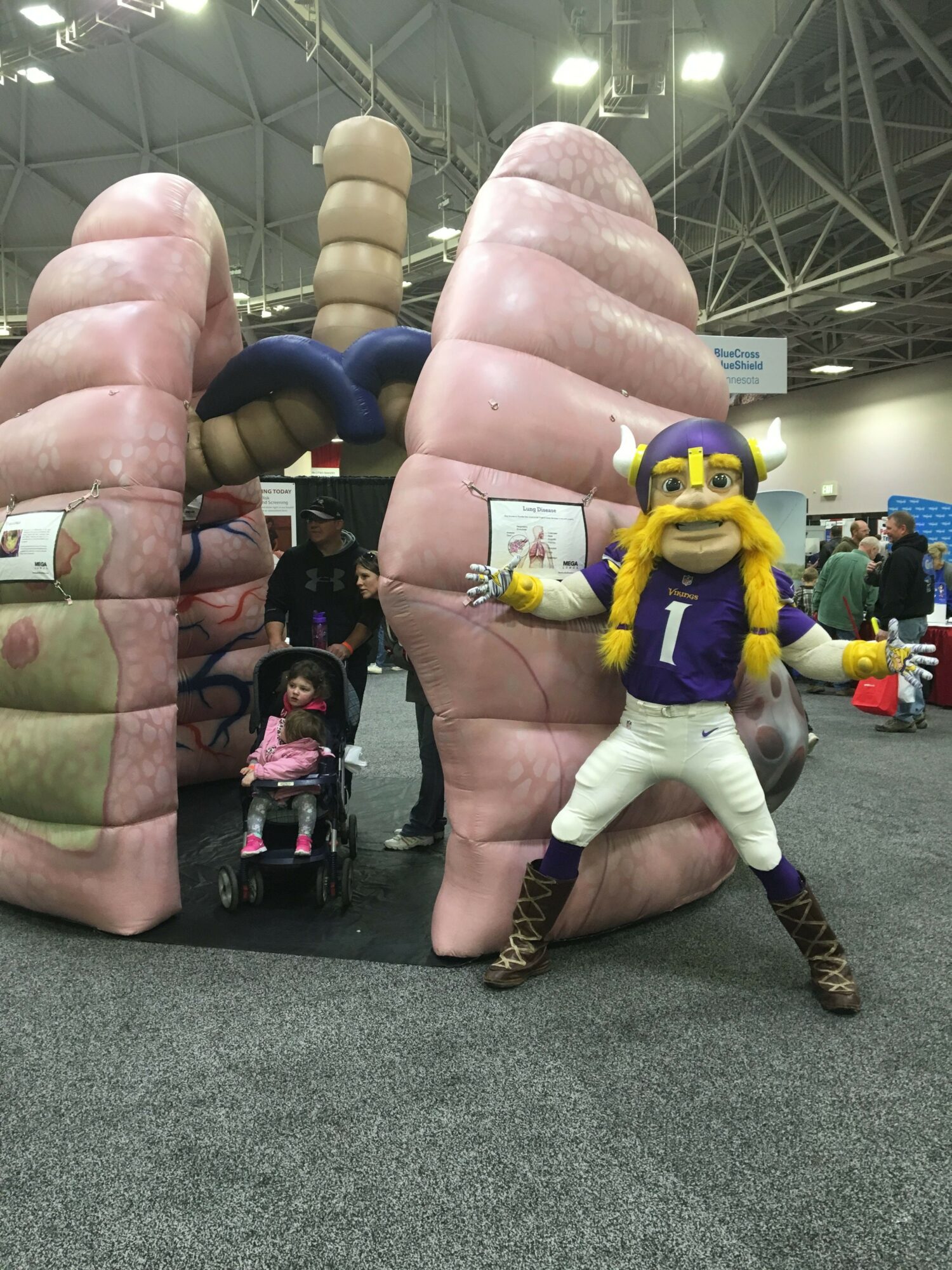 Person in football gear sitting on a large pink sculpture at an indoor event, with a child and a stroller nearby.