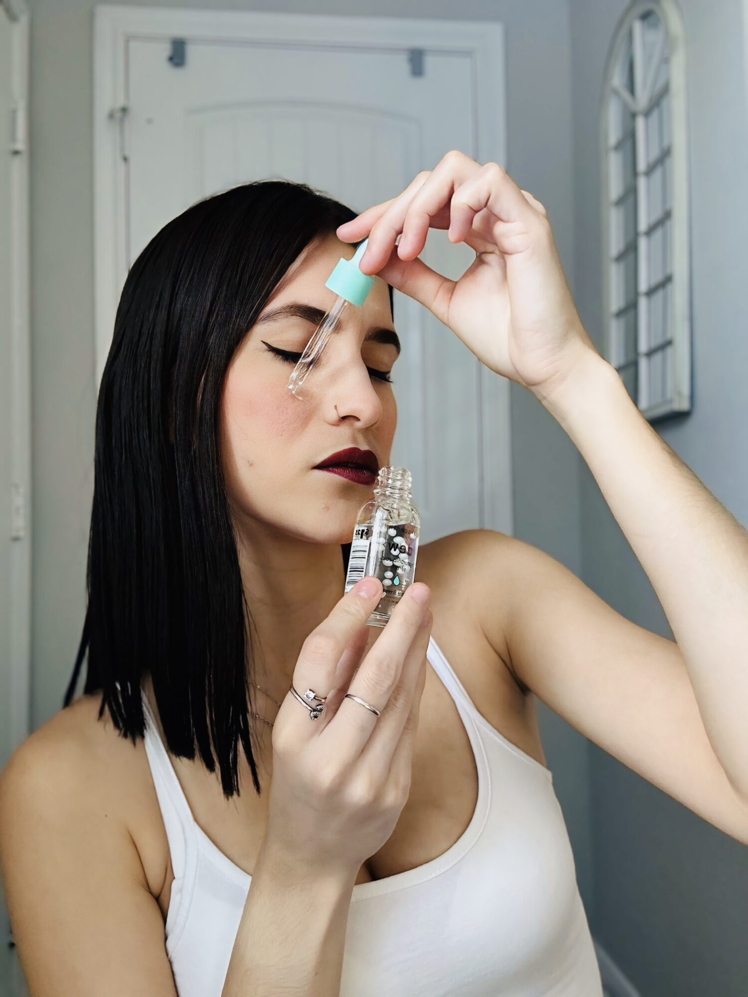 Woman with black hair and dark lipstick applies makeup with a small mirror, wearing a white tank top.