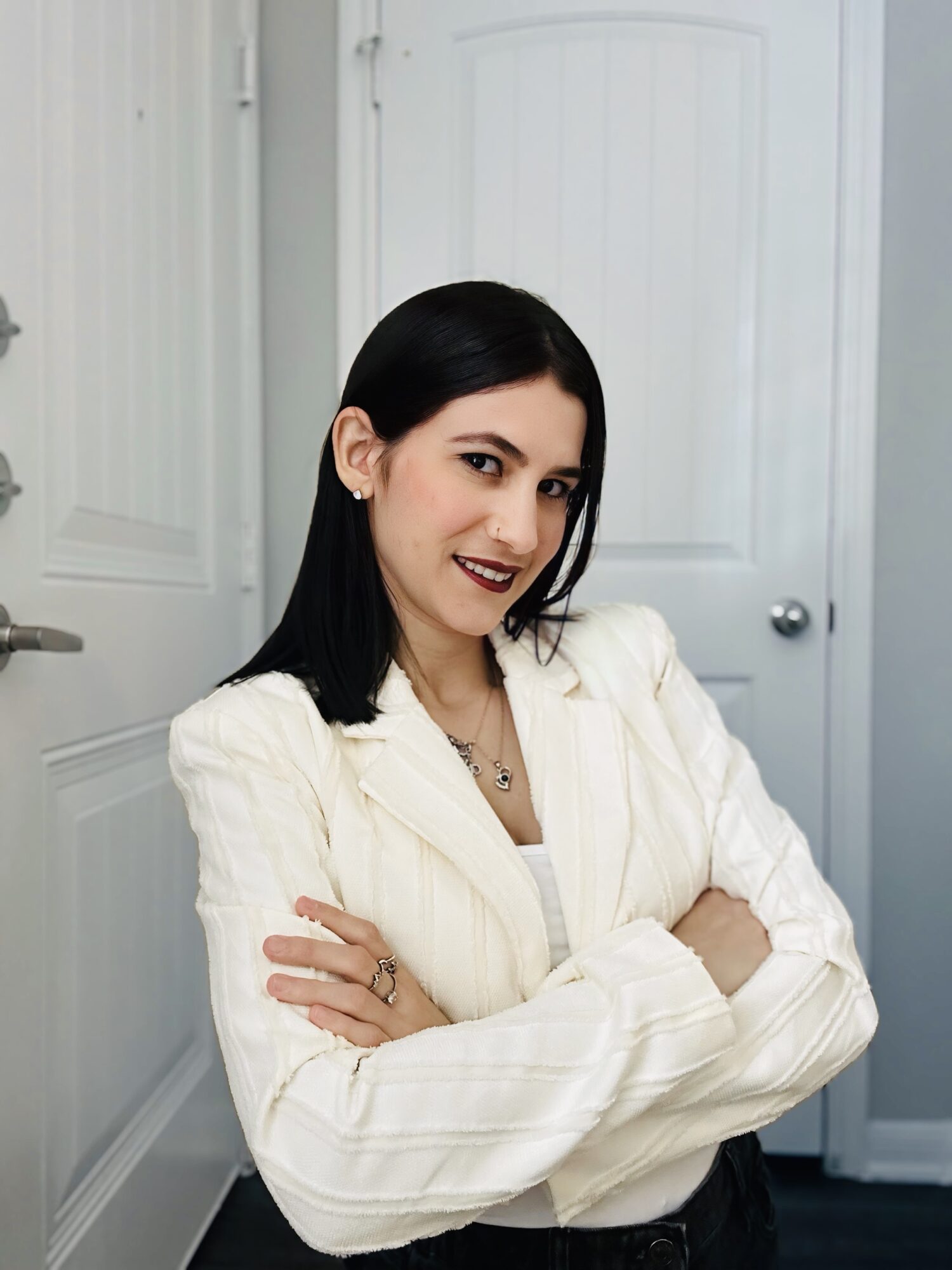 Young woman with black hair and dark lipstick, wearing a white blazer, standing with arms crossed in a room with white walls and door