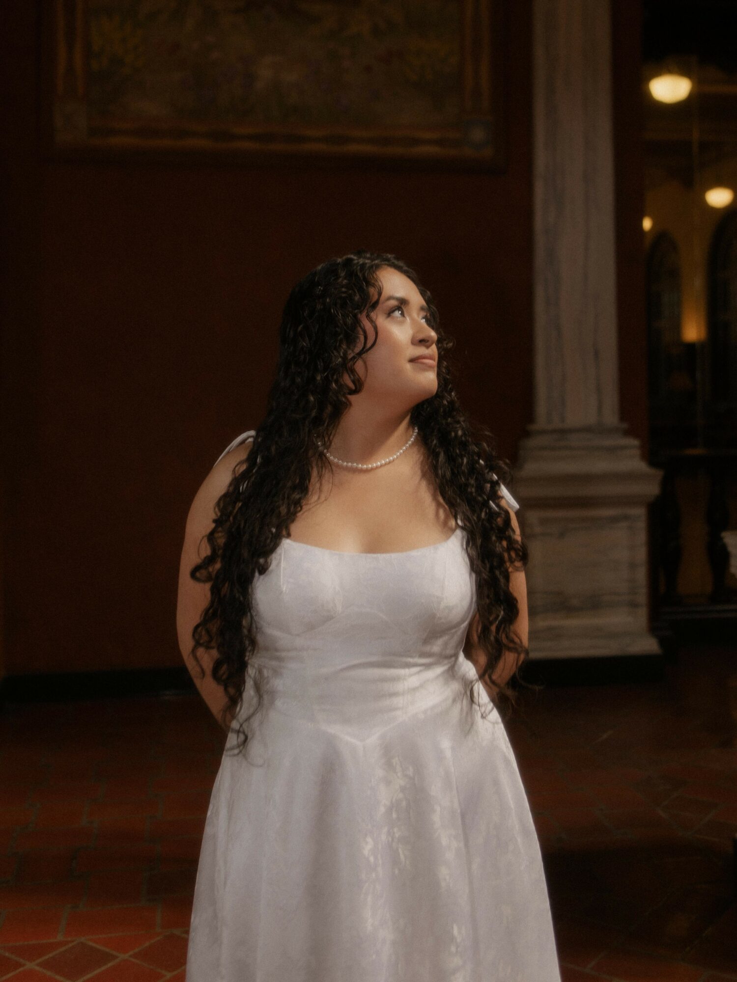 Woman with long curly hair in a white dress standing indoors, looking upward, with warm lighting and architectural details behind her.