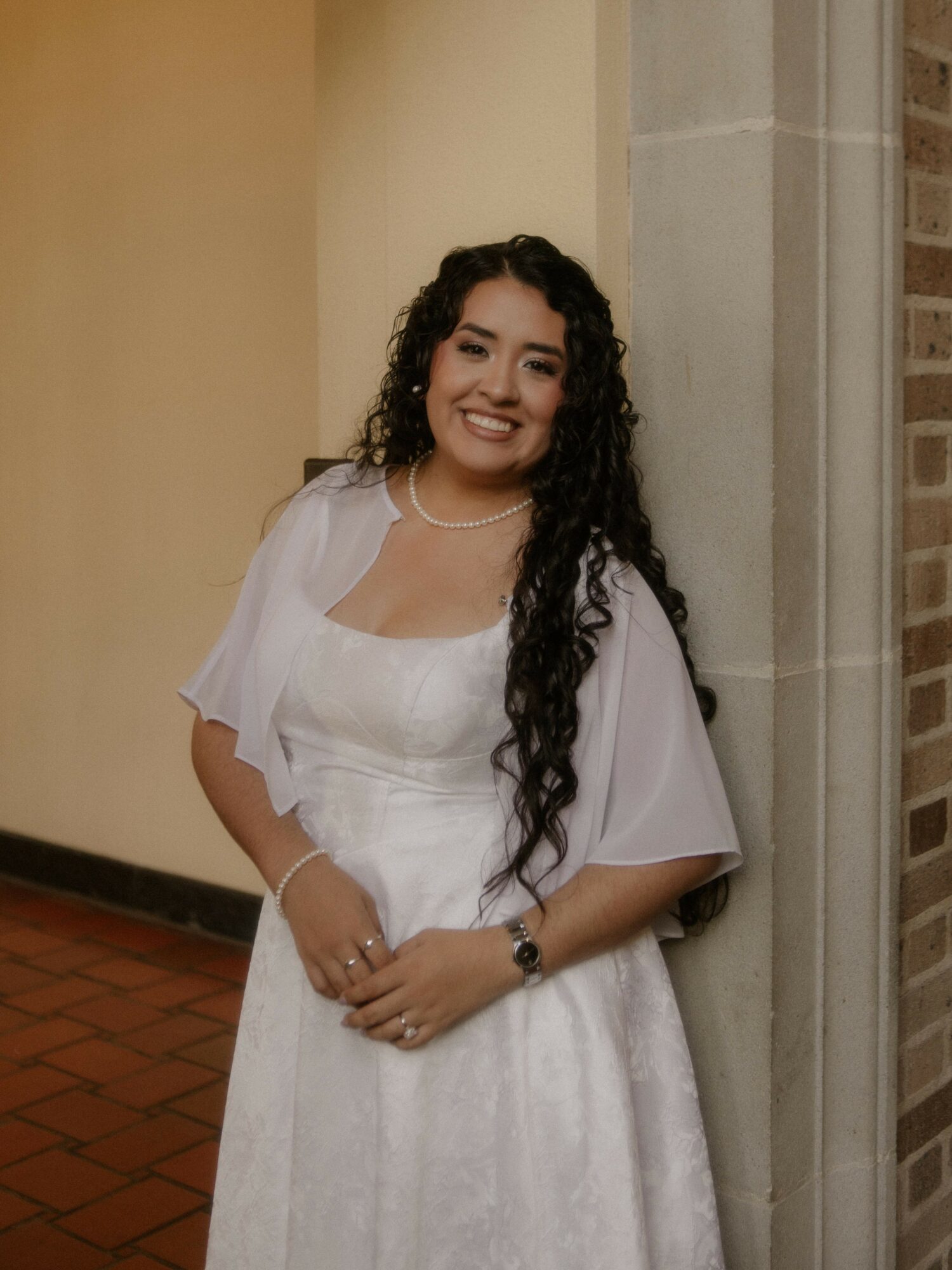 Smiling woman with long curly hair in white dress leaning against a wall, indoors.