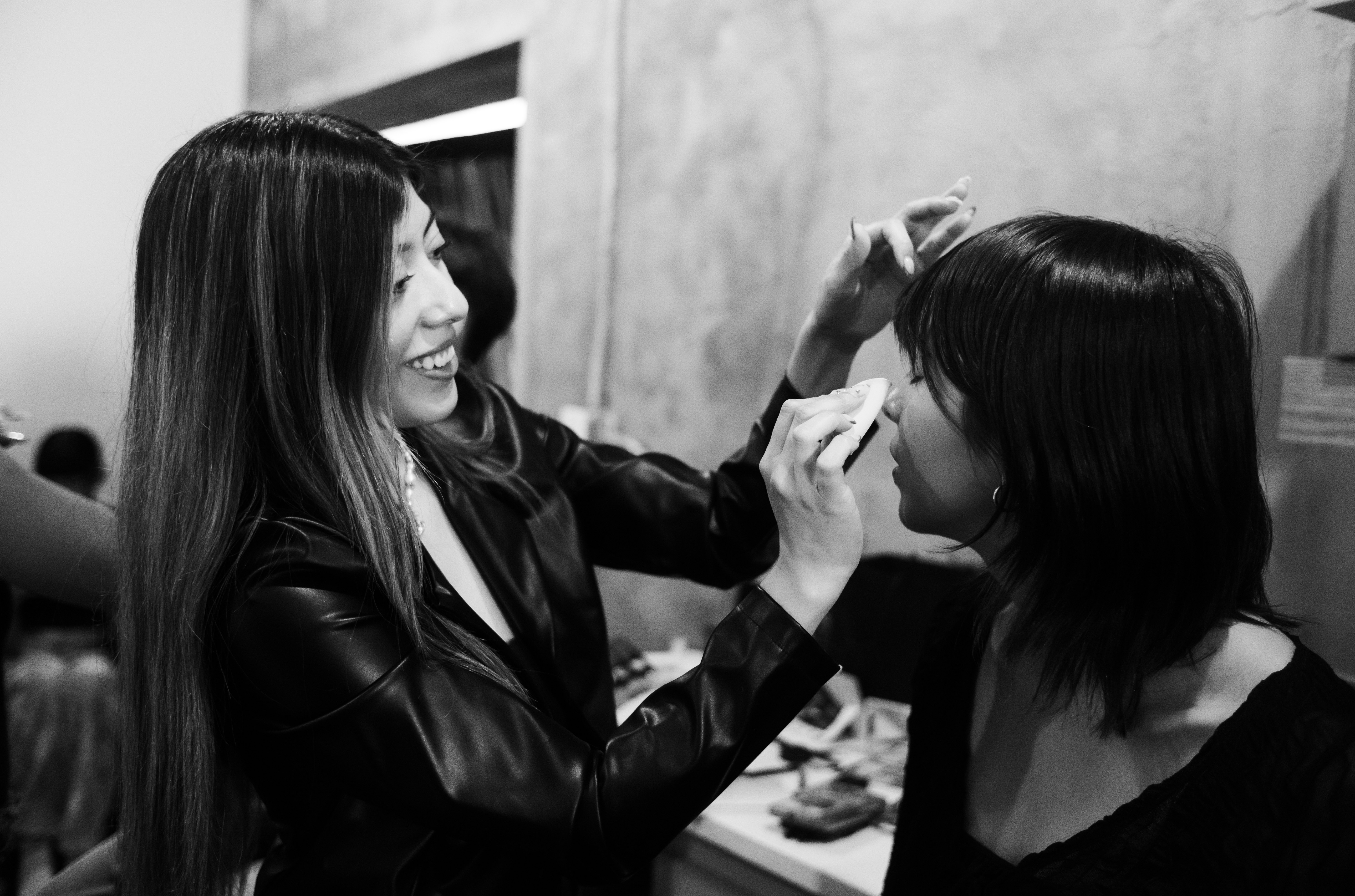 Woman applying makeup to another woman with a smile, in black and white, indoors.