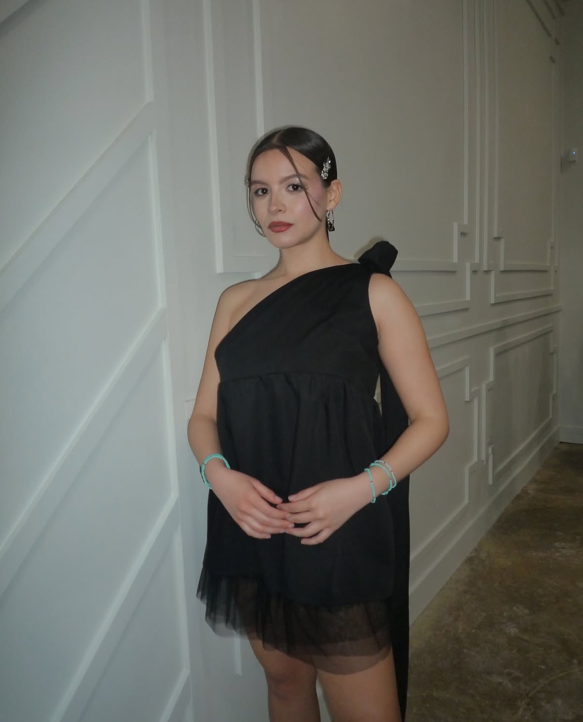 Young woman in black dress standing indoors with white paneled walls, hands clasped, looking at camera.