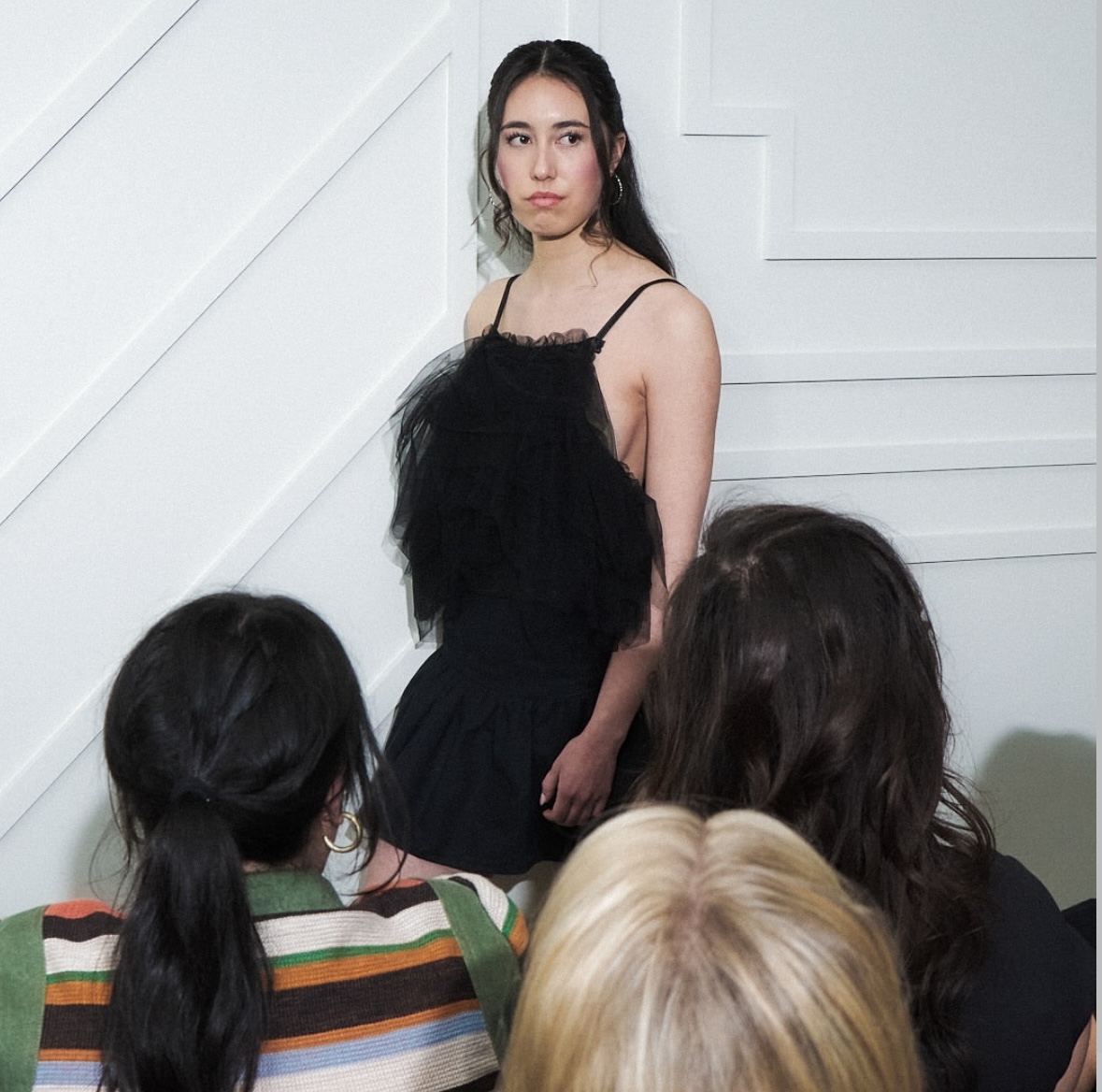 Young woman with long dark hair in a black dress standing in front of a group of people indoors, white wall background.