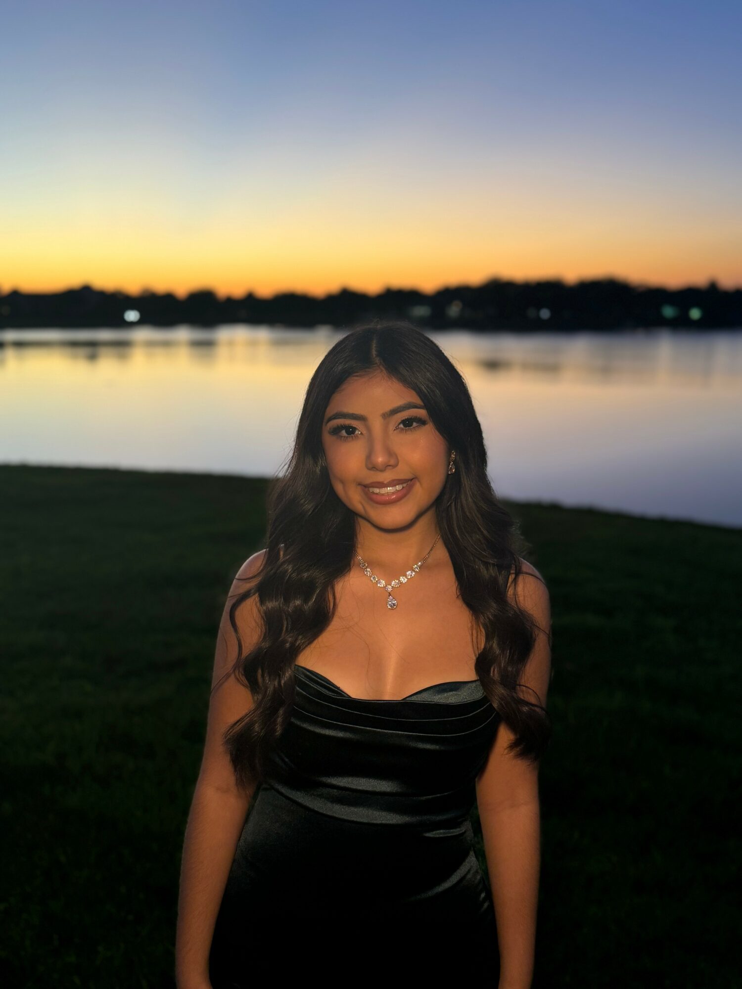 Young woman with long dark hair smiling outdoors at sunset near a body of water.