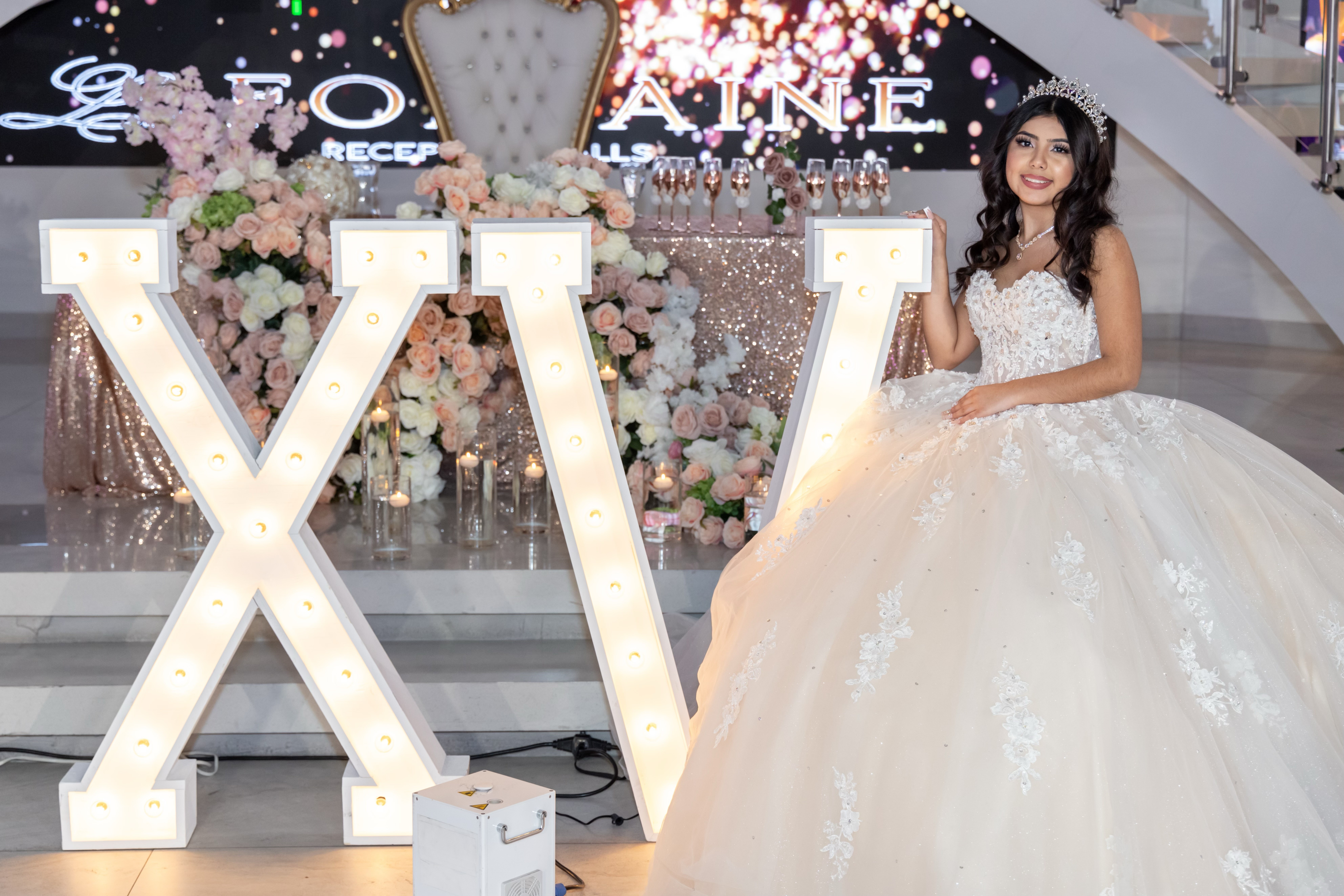 Young woman in a white wedding gown sitting beside large illuminated letters 'X' and 'V' with floral decorations in background.