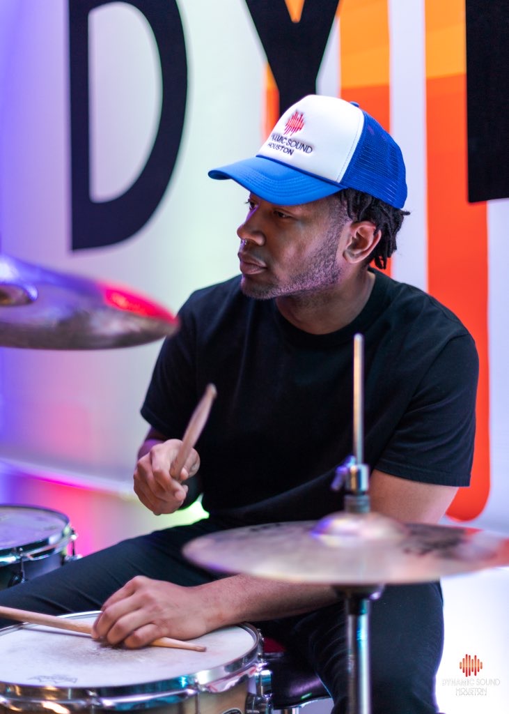 Person playing drums, wearing a white and blue cap, black shirt, in a colorful room with large letters on the wall.