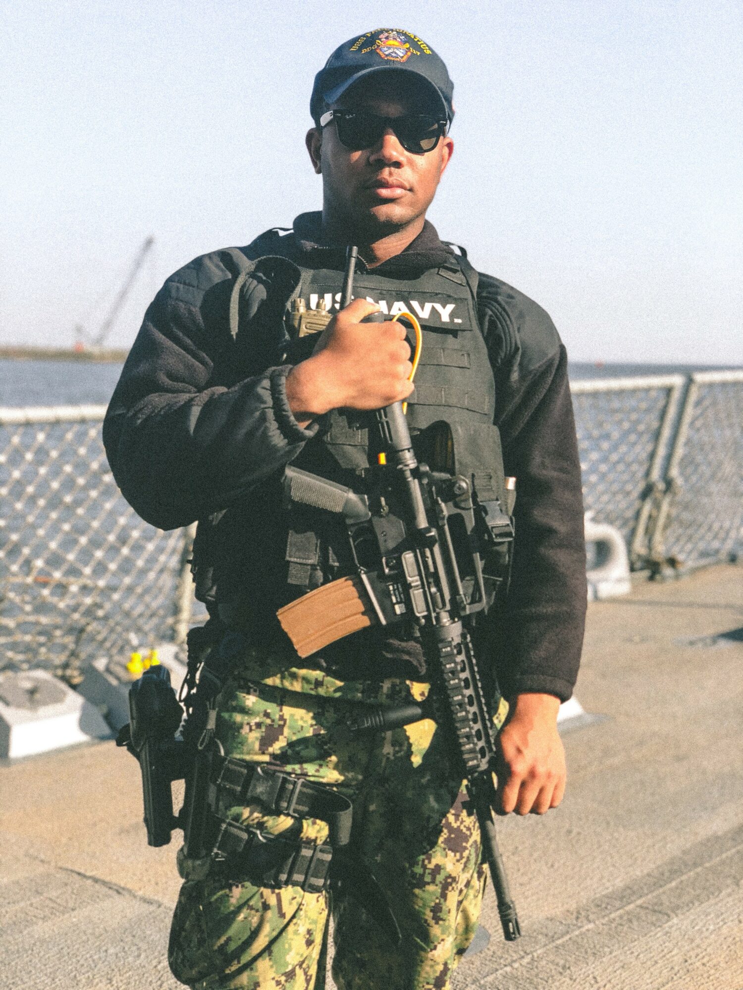 Person in military gear holding a rifle, wearing sunglasses and a cap, standing outdoors near a fence.