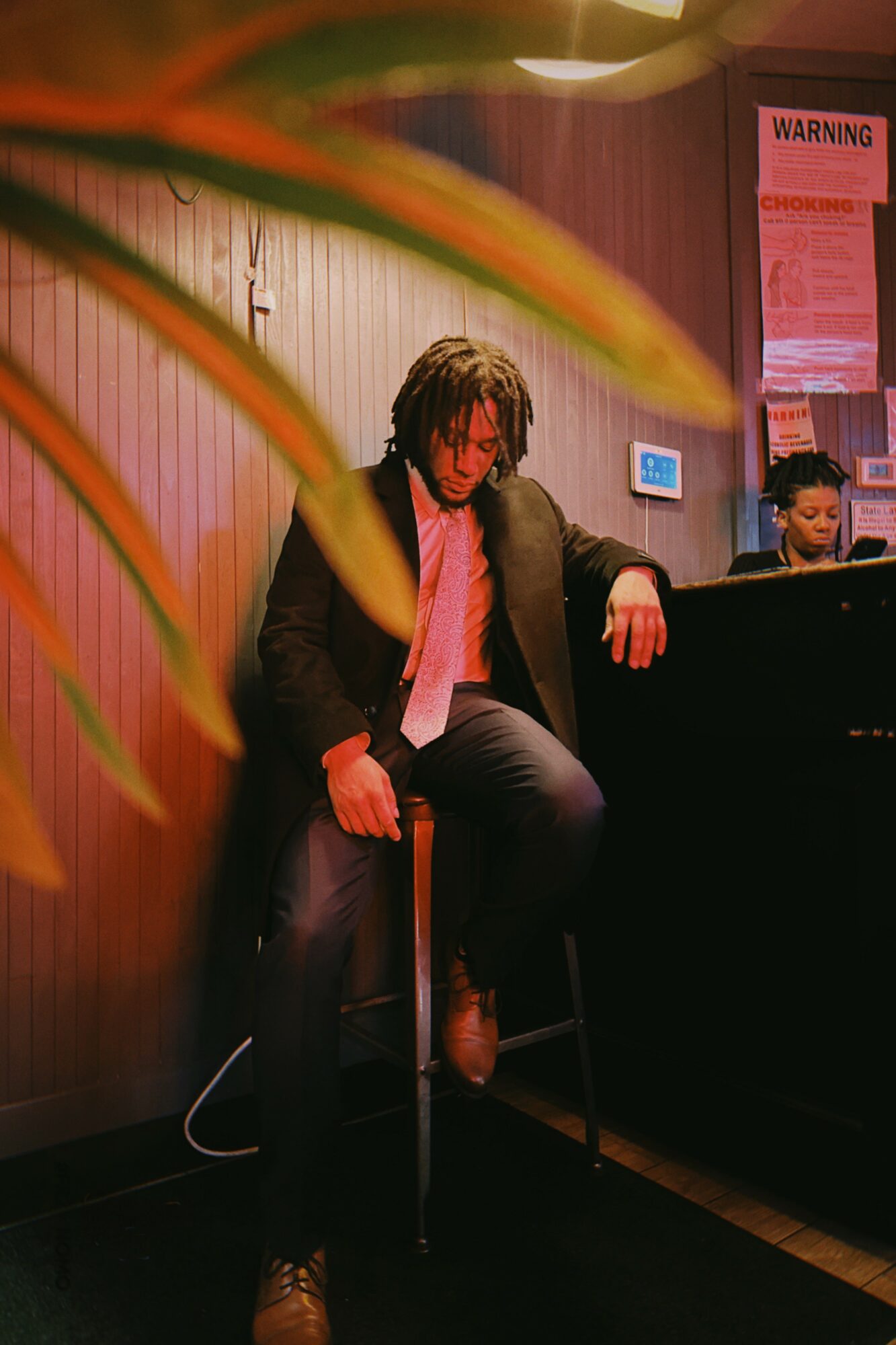 Person with dreadlocks sitting on a stool, playing a piano in a dimly lit room, with a plant partially visible in the foreground.