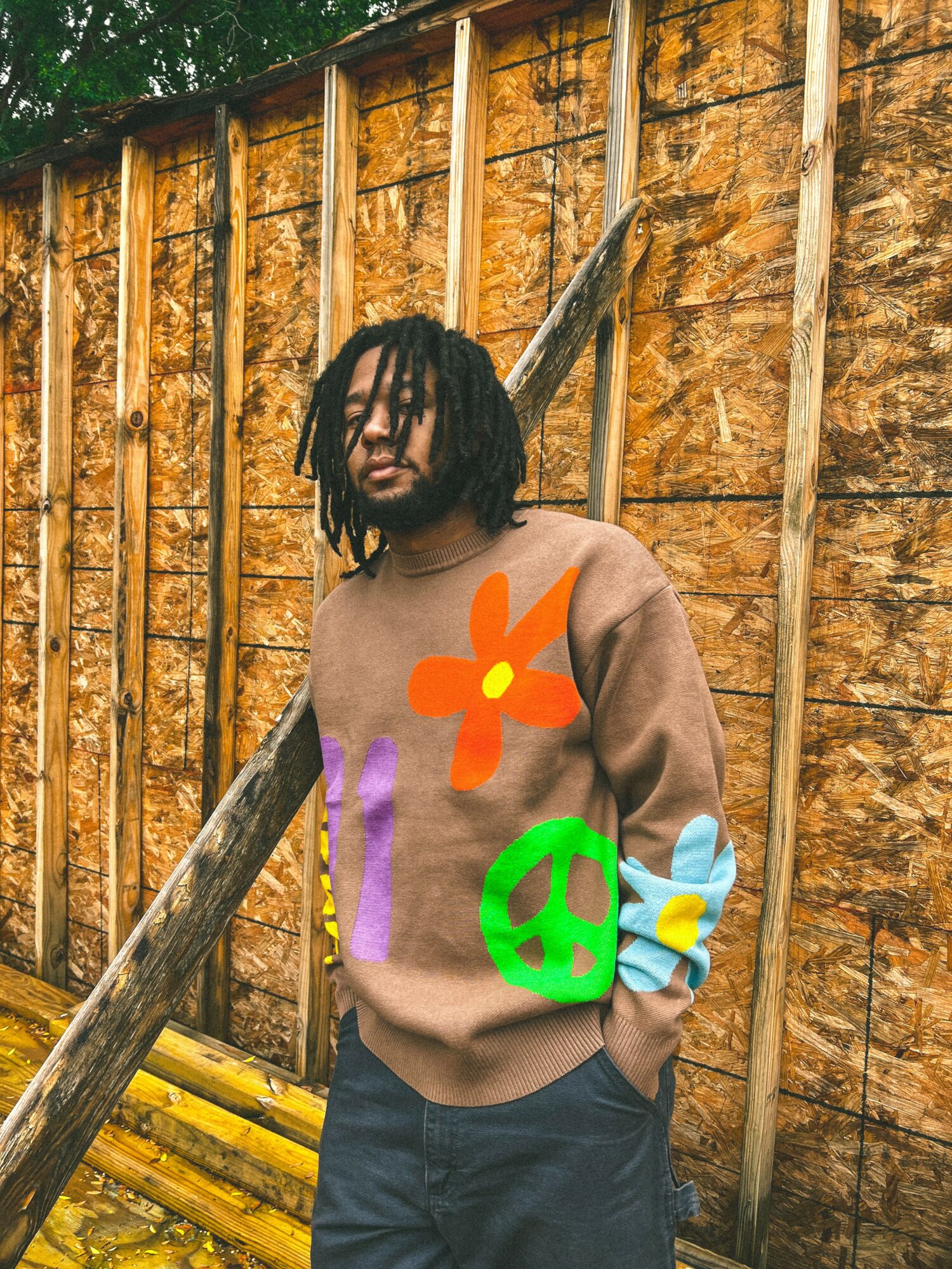 Young person with dreadlocks standing outdoors against a wooden fence, wearing a colorful sweatshirt with peace sign and flower designs.