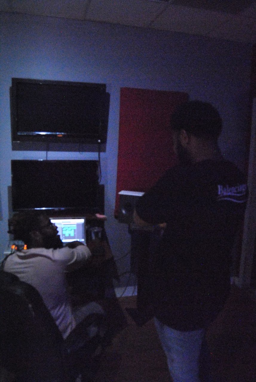 Two people in a dimly lit room with a TV and a computer, one sitting and one standing, facing away from the camera.