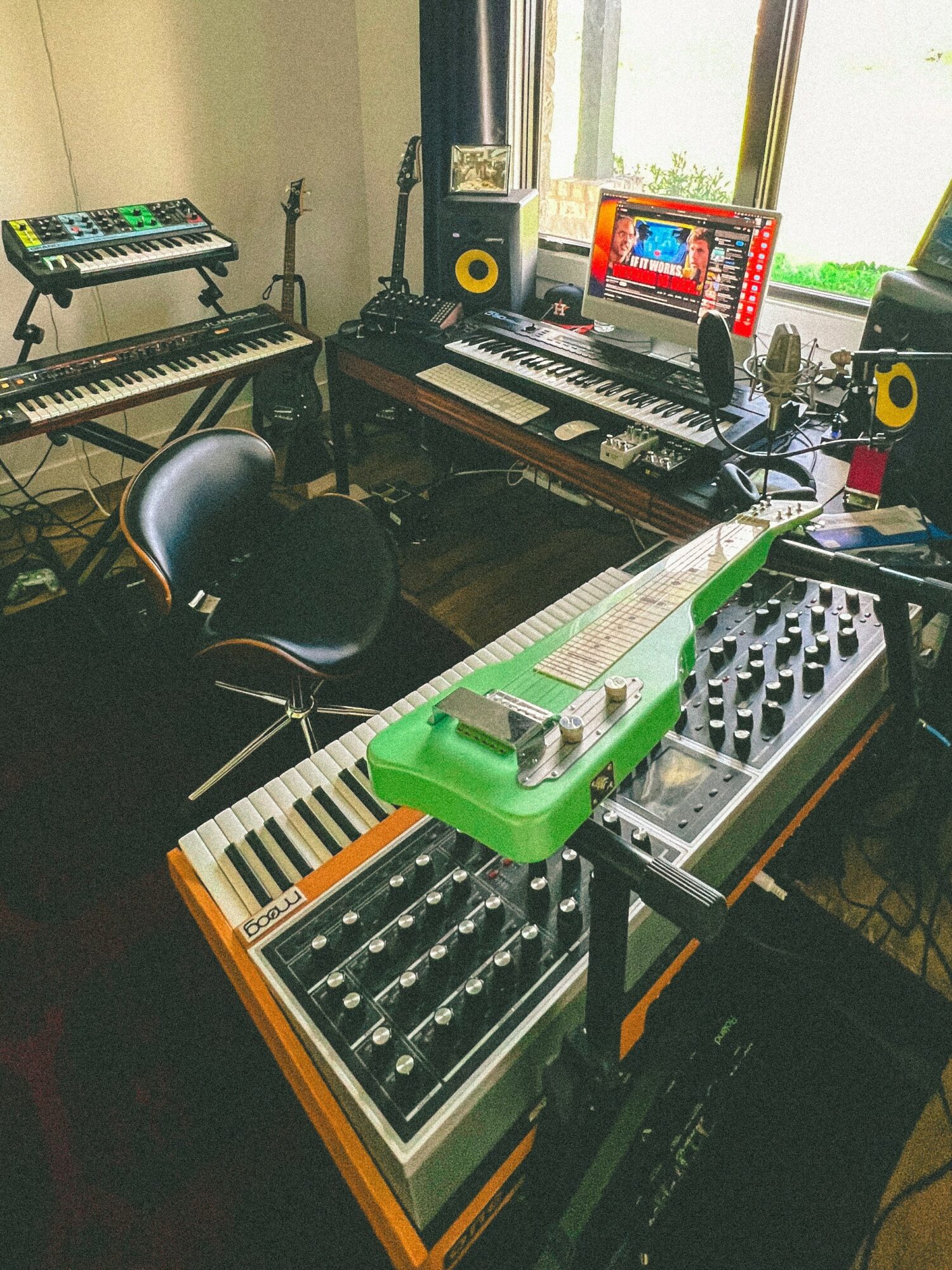 Music studio with keyboards, synthesizers, and a computer on a desk near a window, with a chair and audio equipment.