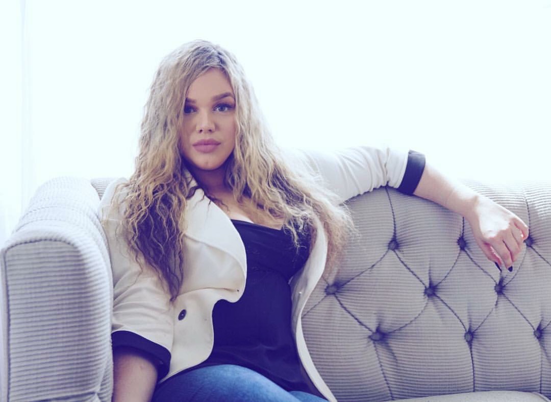 Woman with long curly hair sitting on a tufted sofa, wearing a white jacket and black top, in a bright room.
