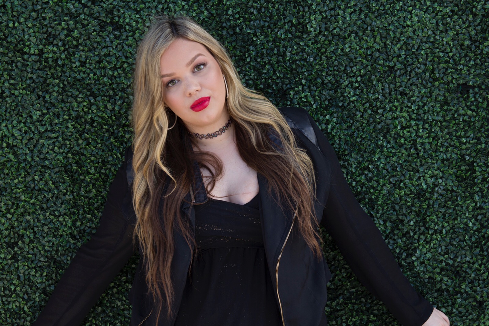 Young woman with long wavy hair, red lipstick, black top, and black jacket, standing against a green hedge background.