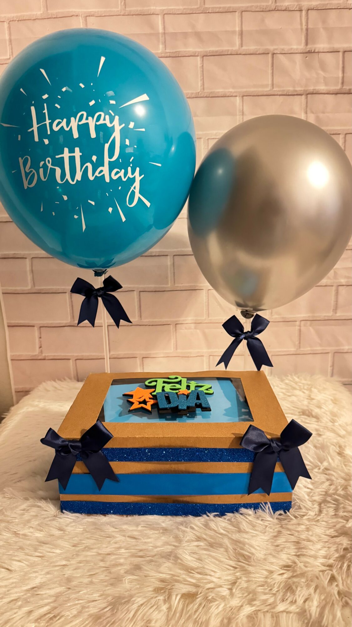 Two balloons, one blue with 'Happy Birthday' written, and one silver, with a birthday cake on a table with blue ribbons.