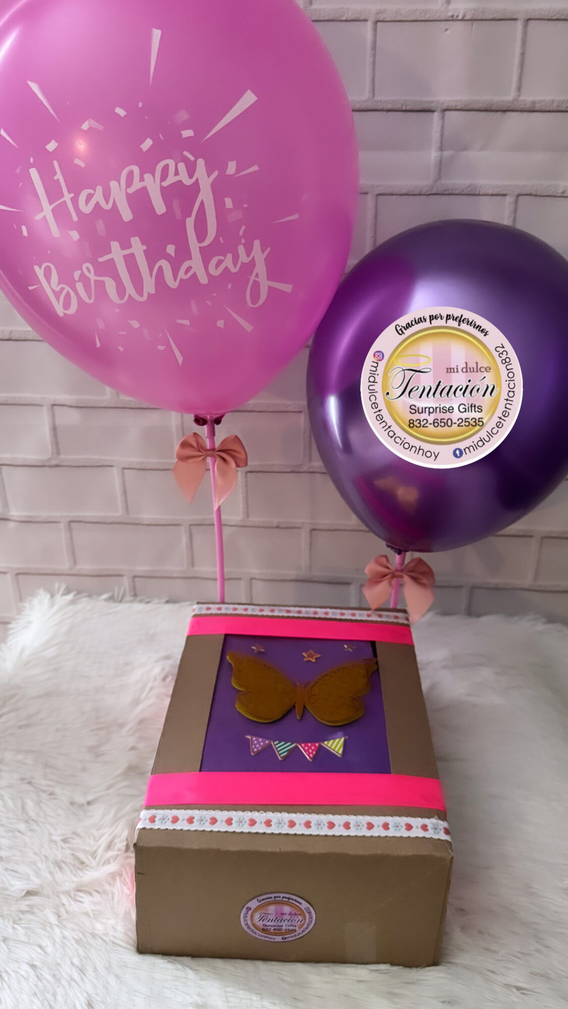 Pink and purple balloons with birthday messages and a gift box with a bow, set against a brick wall.