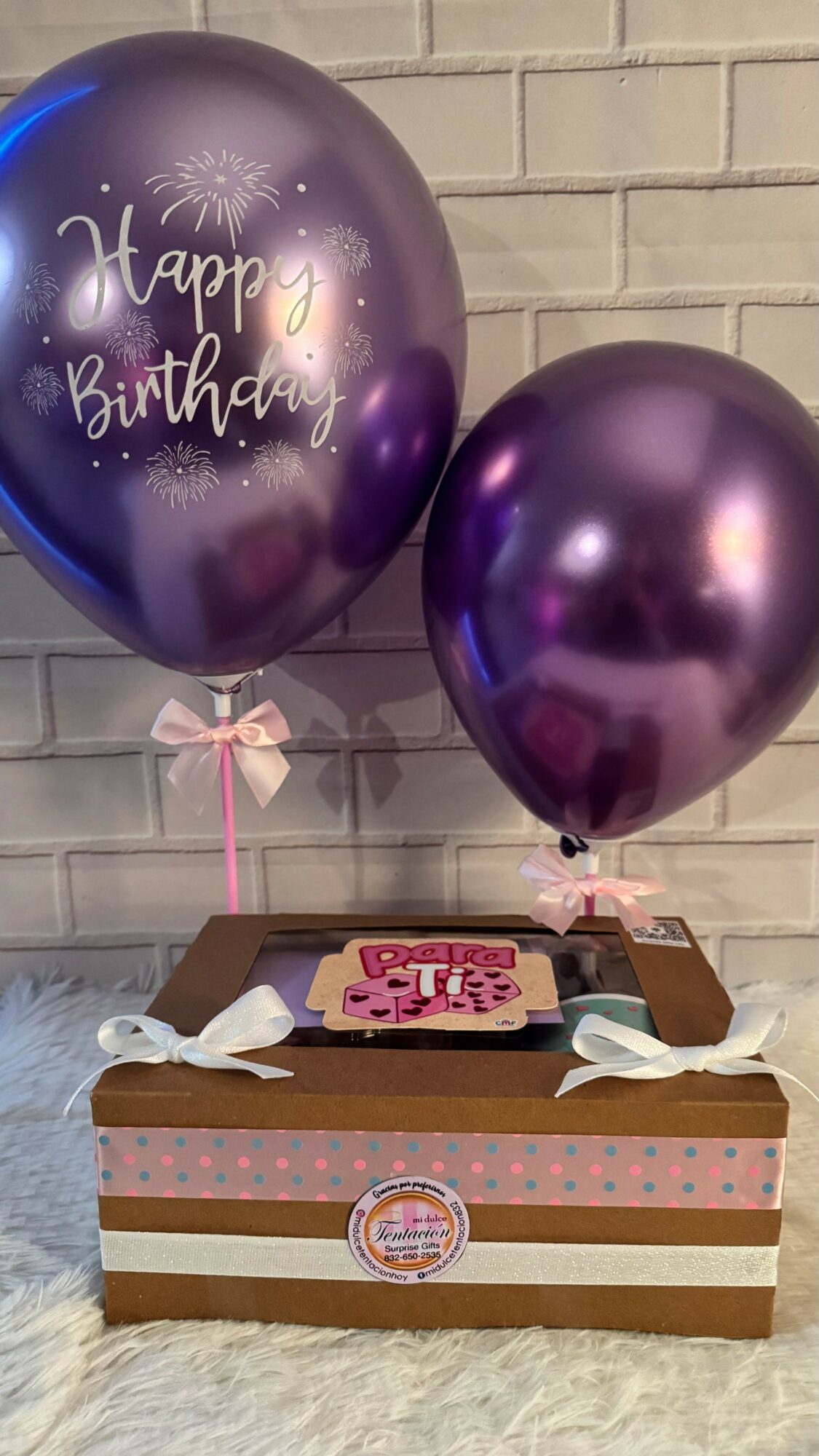 Two purple balloons, one with 'Happy Birthday' text, attached to a decorated gift box against a brick wall.