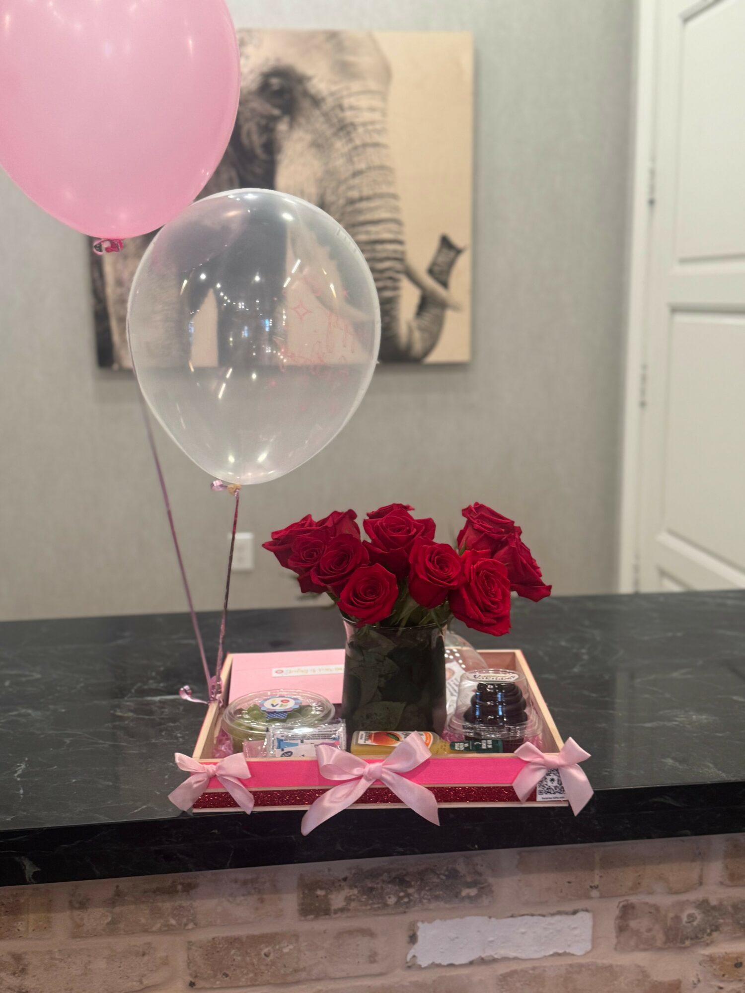 Pink and clear balloons, red roses in a vase, and small gifts on a tray with pink ribbons on a dark table.