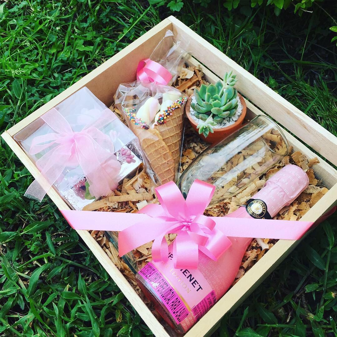 Open gift box with pink ribbon, containing a small potted succulent, a watch, and wrapped items, on grass.