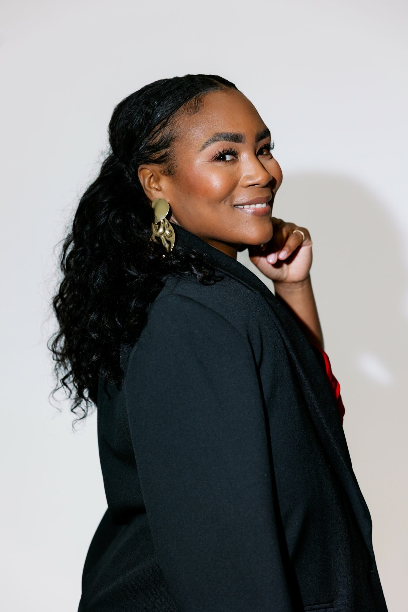 Smiling woman with long curly hair, wearing earrings and a dark blazer, posing against a plain background.