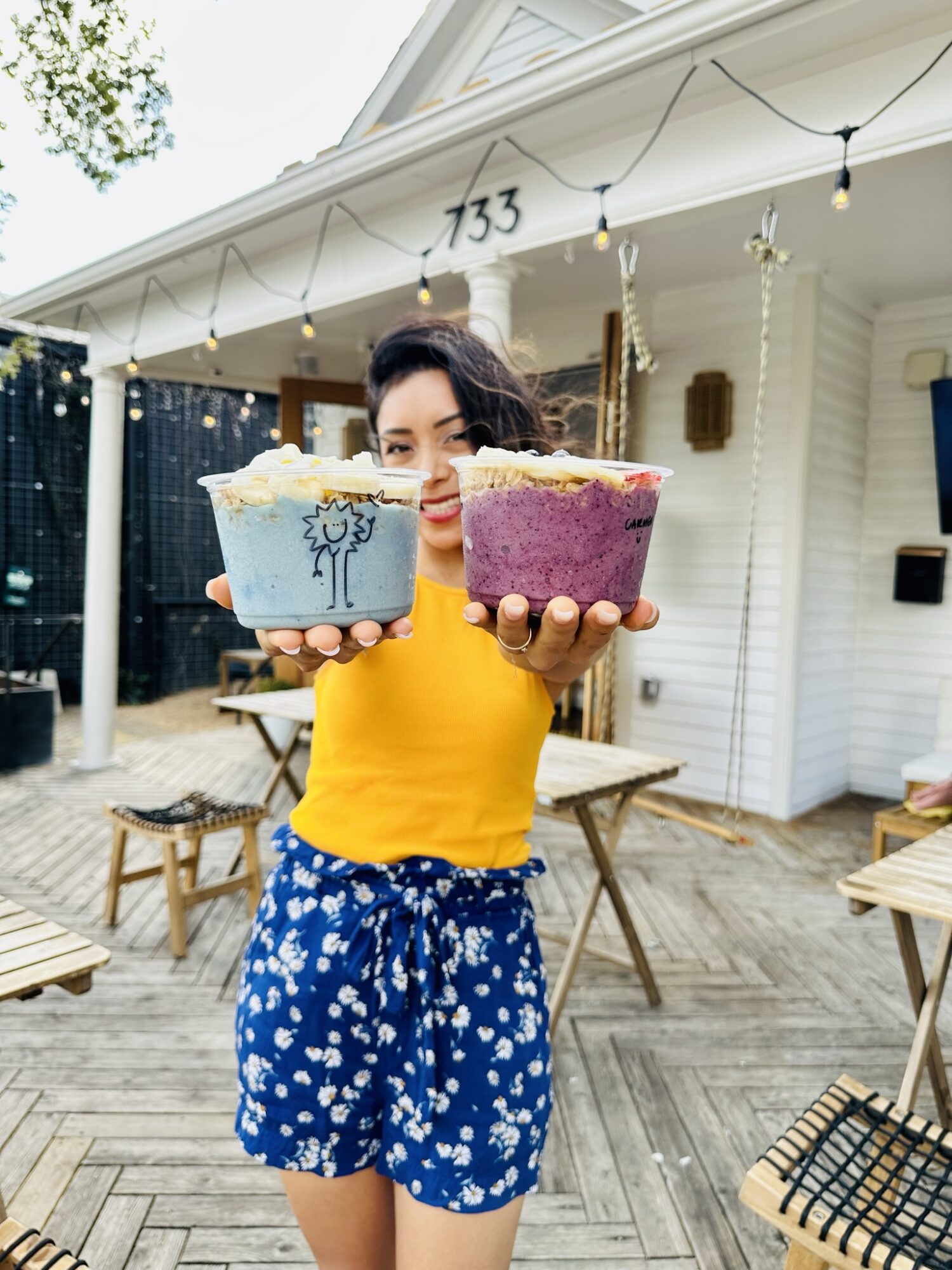 Person holding two colorful cakes outdoors in front of a white house with string lights.