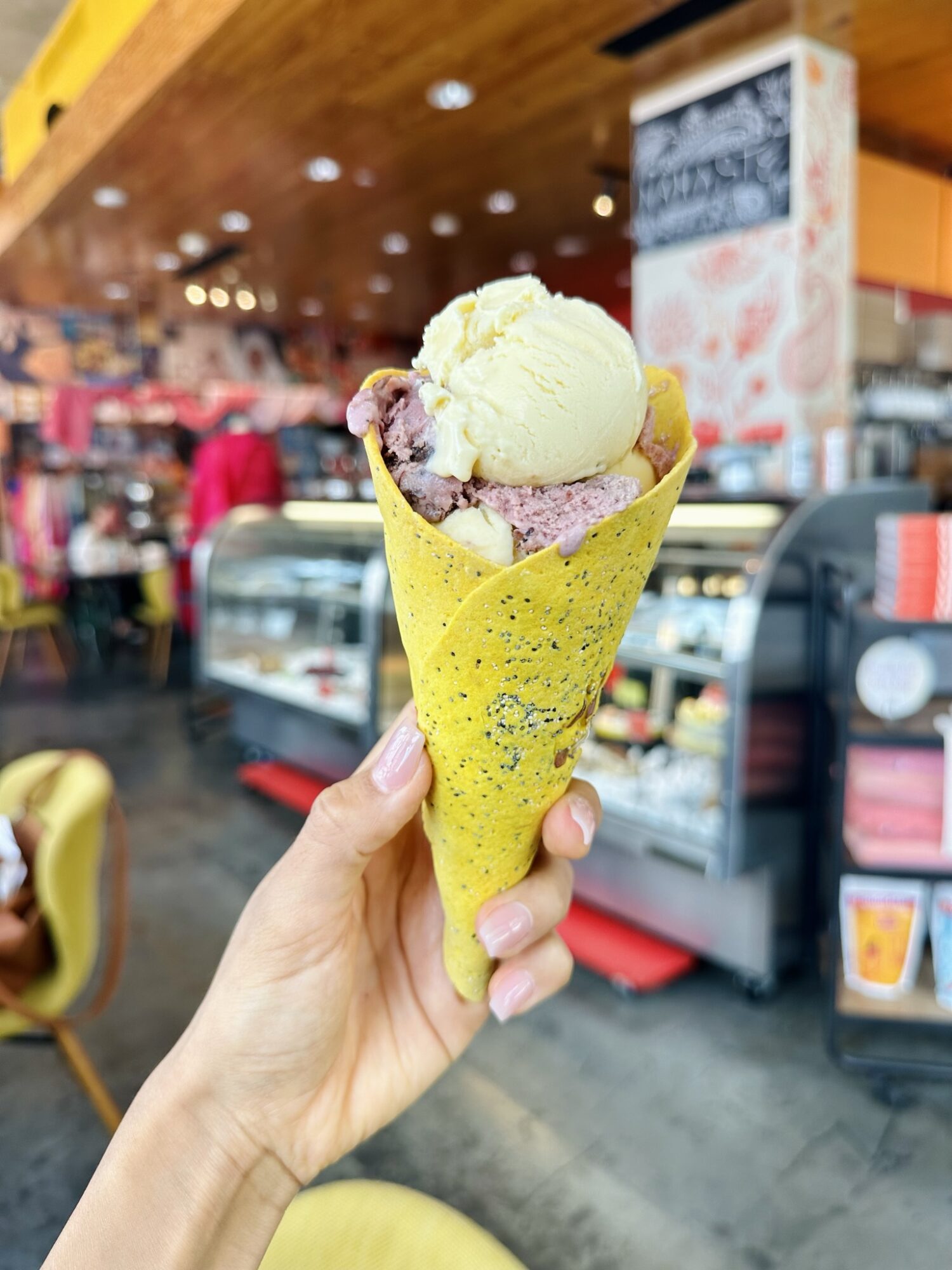 Hand holding a yellow cone with ice cream and toppings inside a cafe, blurred background of cafe interior.
