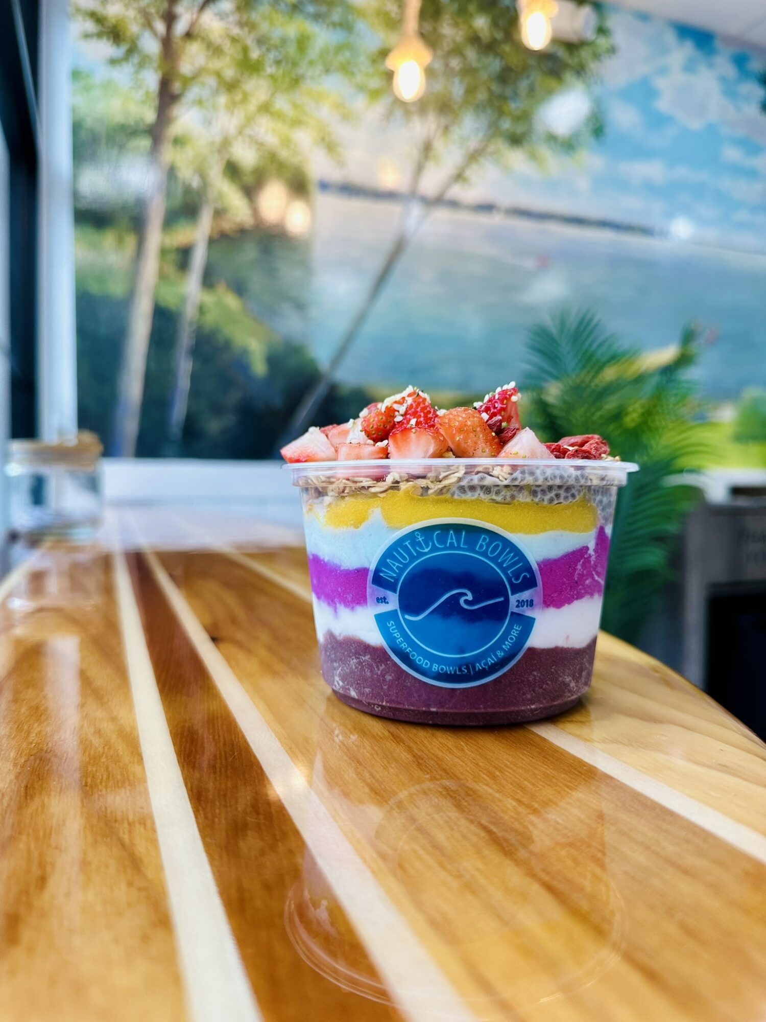Colorful layered dessert in a clear cup with strawberries on top, placed on a wooden table with a blurred outdoor background.