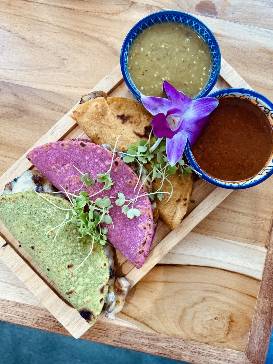 Three colorful Indian-style flatbreads with microgreens, served with two bowls of chutney and sauce on a wooden tray.