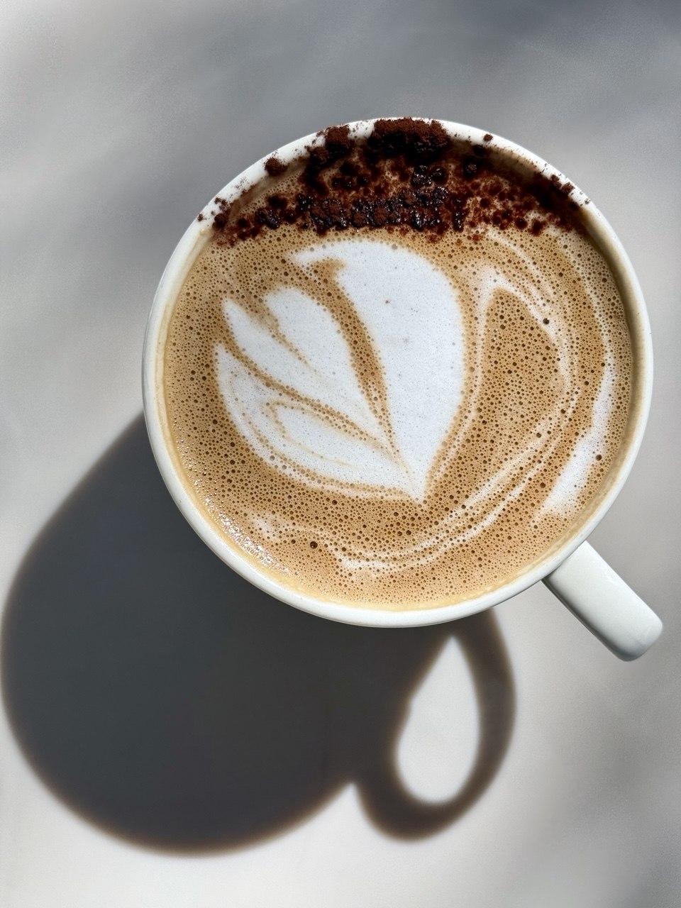 Top-down view of a coffee cup with latte art, shadow cast on surface, and a handle visible on the right.