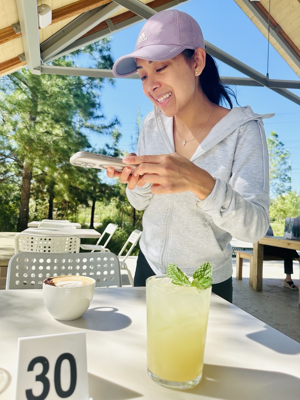 Young woman in a light hoodie and cap smiling while looking at her phone outdoors, with a drink and bowl on table.